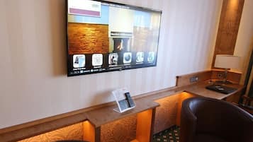 A hotel reception area with a flat-screen TV displaying a welcome message, a reception desk, two leather chairs, and a small table with a remote control.