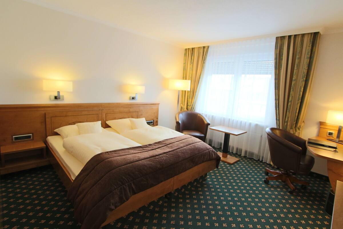 A hotel room with a large bed, two bedside lamps, a desk with a chair, and a window with curtains.
