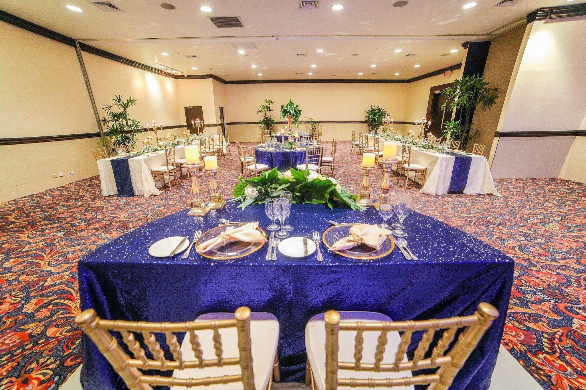 Un salón de banquetes con mesas redondas cubiertas con manteles azules, sillas doradas y centros de mesa florales.