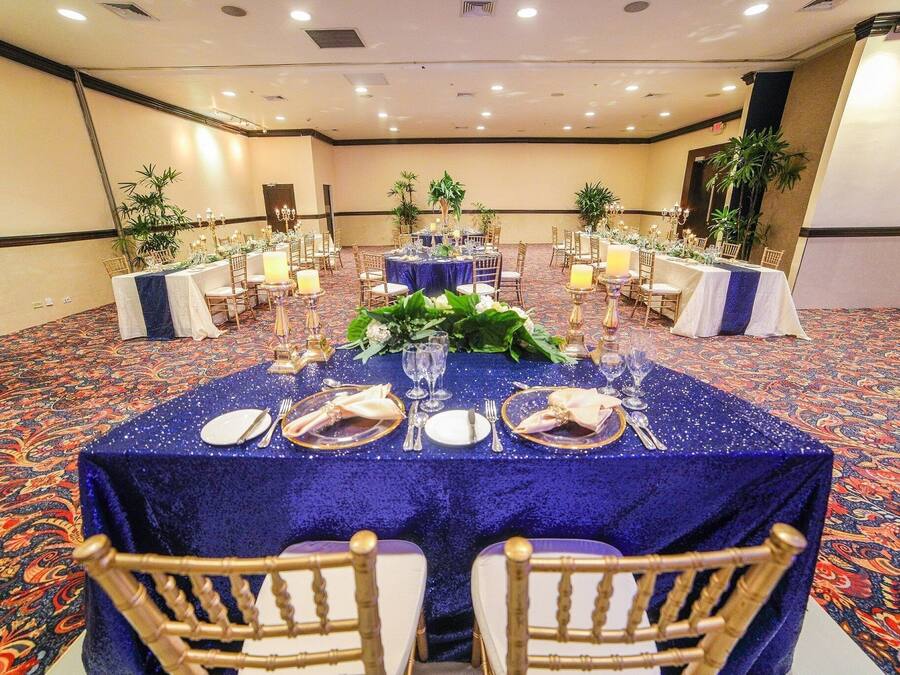 Un salón de banquetes con mesas redondas cubiertas con manteles azules, sillas doradas y centros de mesa florales.