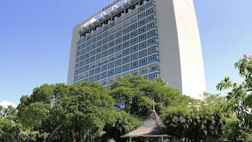 A tall, modern hotel with a white facade and blue accents, surrounded by lush green trees and a small gazebo.