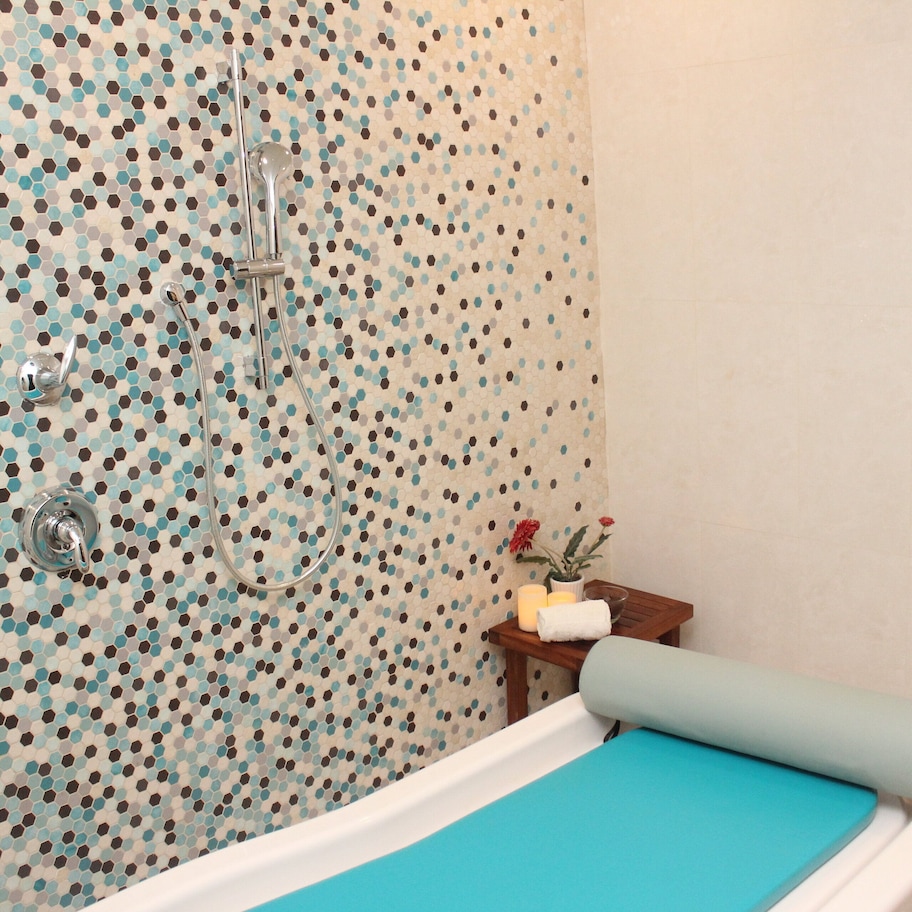 A bathroom with a polka dot tile wall, a showerhead, a small wooden table with toiletries, and a bathtub with a blue mat.