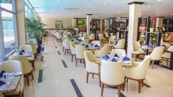 A spacious hotel dining area with multiple tables and chairs, large windows, and potted plants.