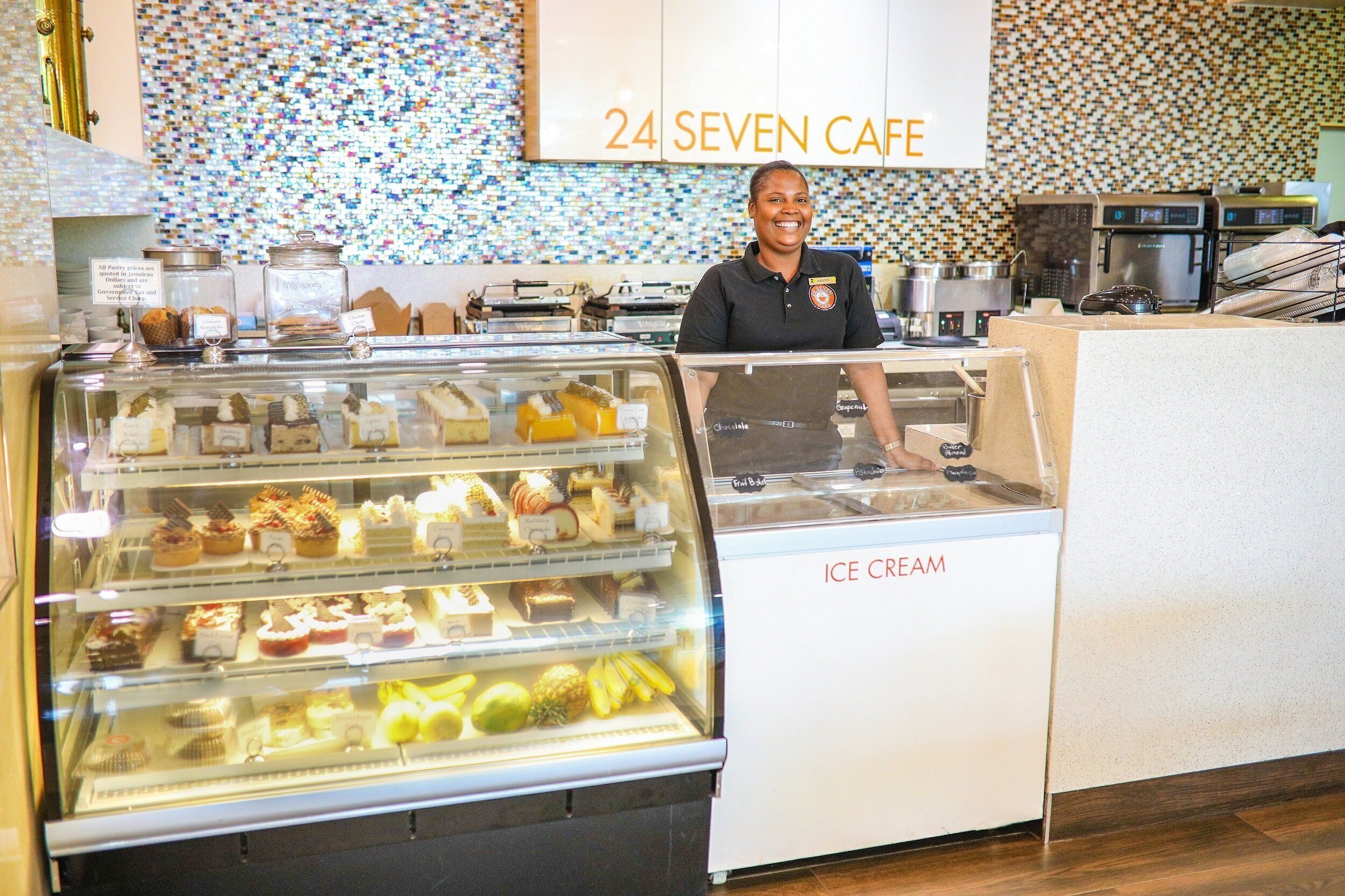 Une personne se tient derrière un présentoir de glaces dans un café, et on peut voir en arrière-plan une enseigne indiquant « 24 SEVEN CAFE ».
