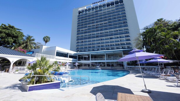 A hotel with a large pool area, multiple lounge chairs, and umbrellas. - The Jamaica Pegasus Hotel (Kingston)