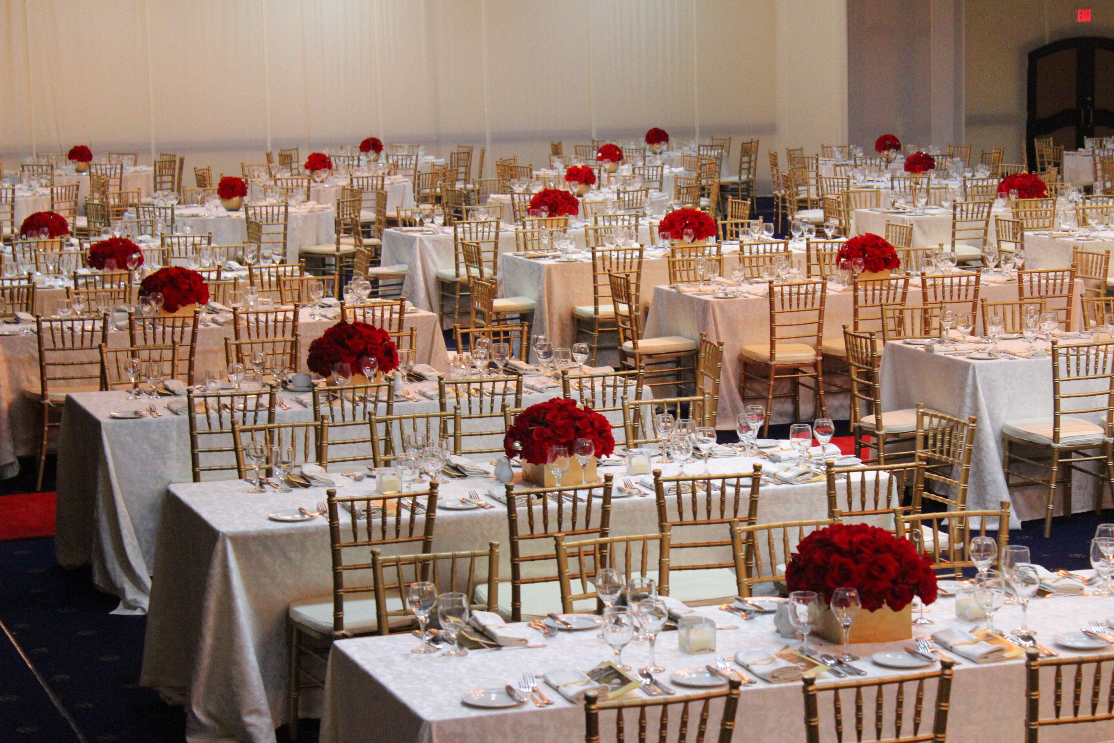 A banquet hall with multiple round tables, each set with white tablecloths, gold chairs, and red floral centerpieces.