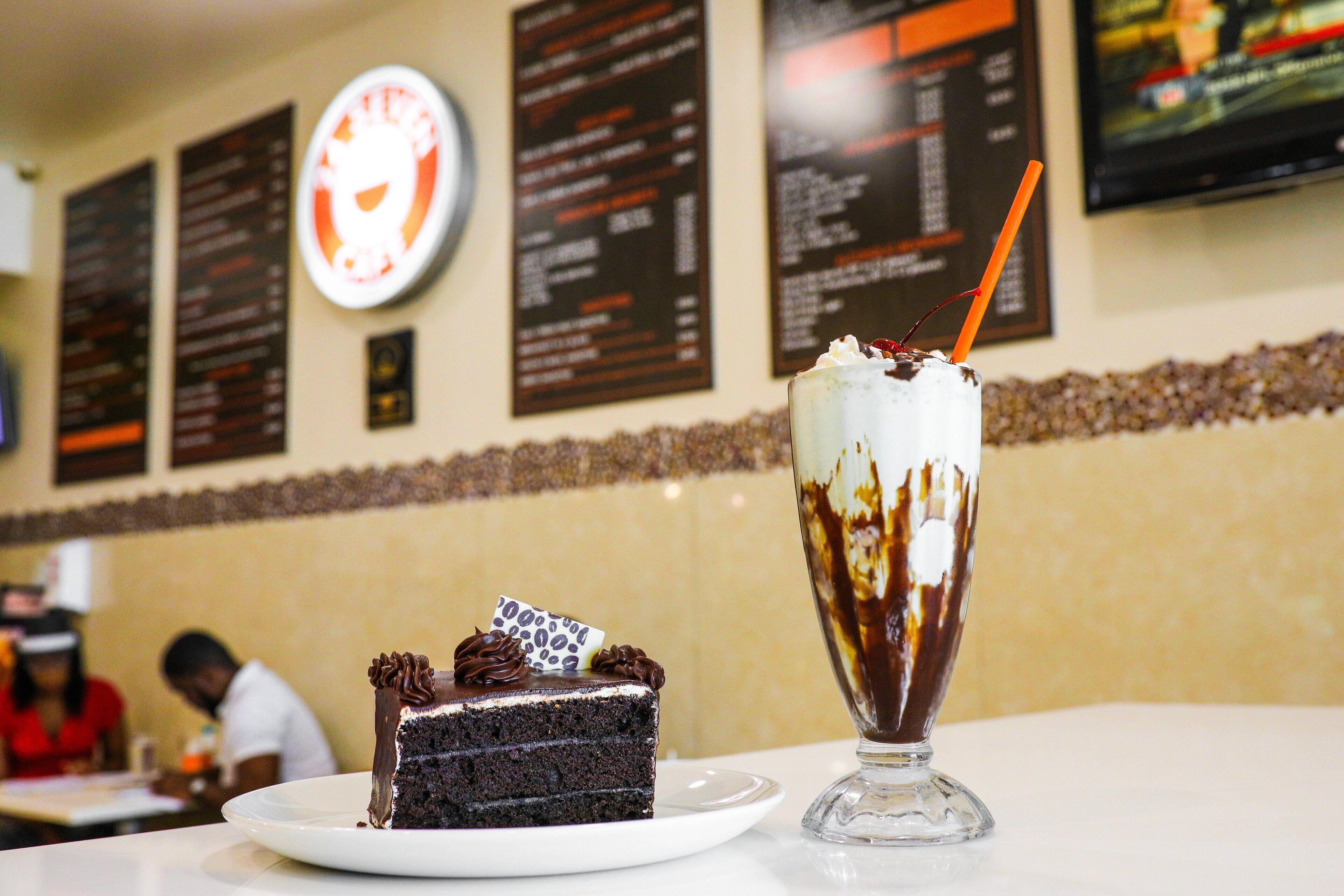 A slice of chocolate cake with whipped cream and chocolate shavings on a white plate, next to a glass of chocolate milkshake with a straw.