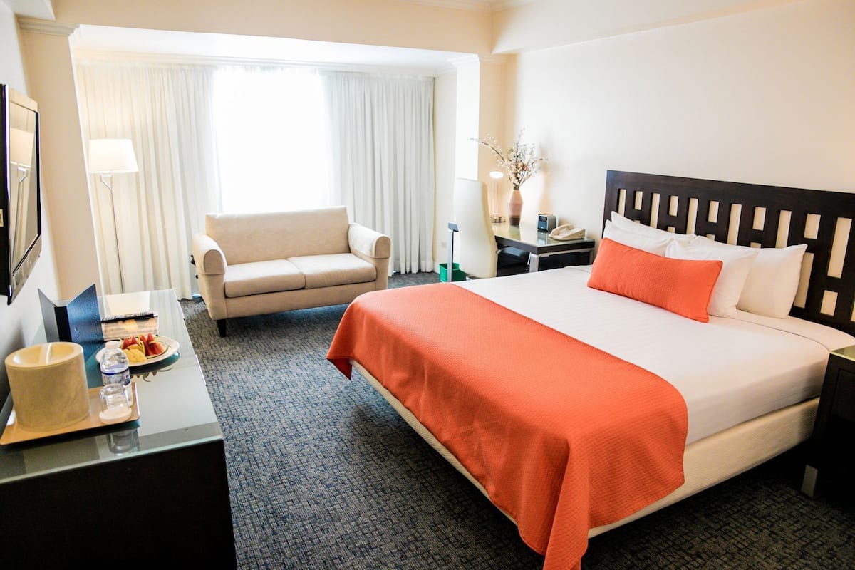 Une chambre d’hôtel avec un lit, une chaise, un bureau avec une lampe, un vase de fleurs et une fenêtre avec des rideaux.