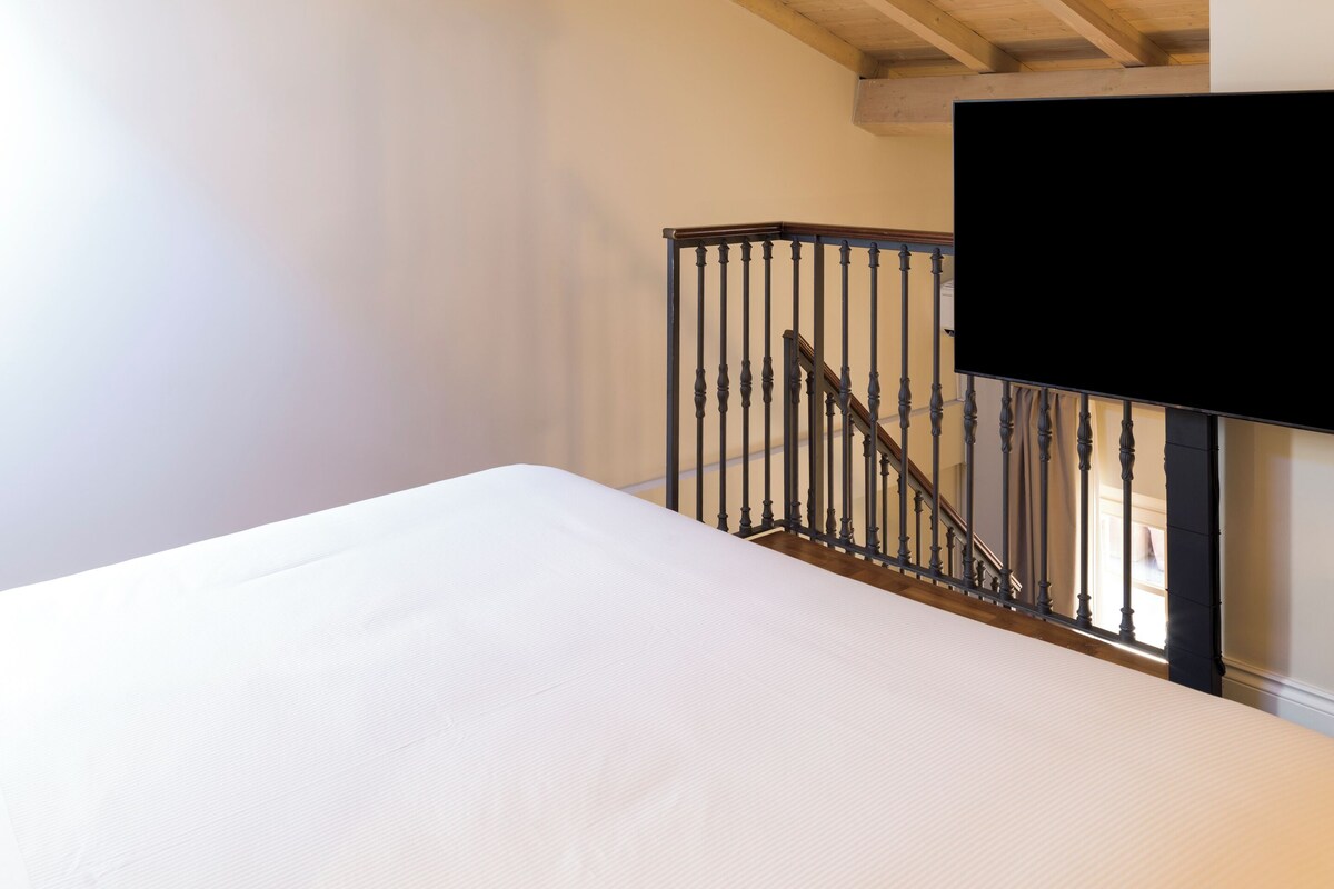 A bedroom with a wooden ceiling, a bed, a staircase, and a TV mounted on the wall.