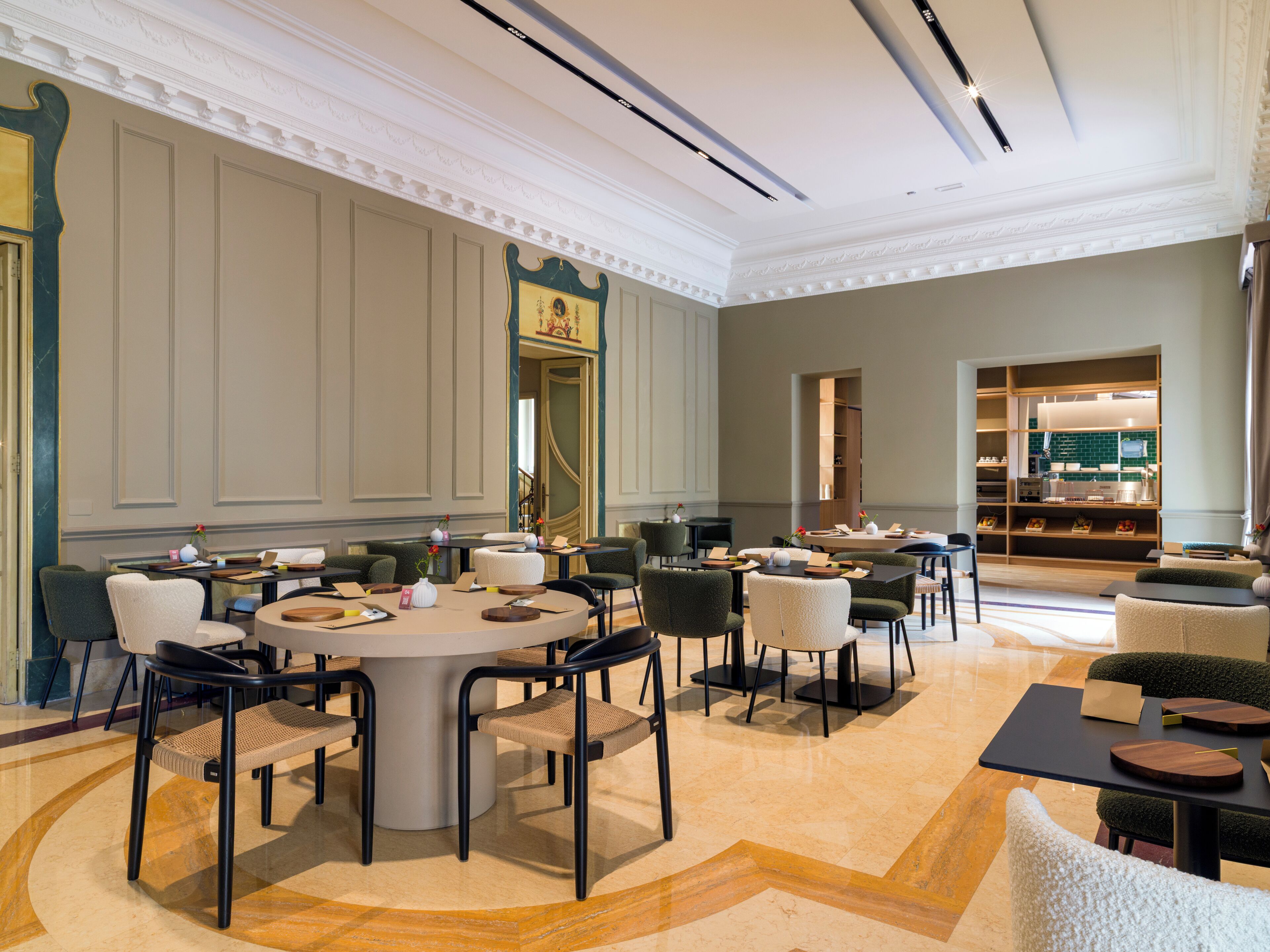 A spacious hotel lobby with a central round table surrounded by chairs, a seating area with sofas, and a kitchen area visible through an open door.