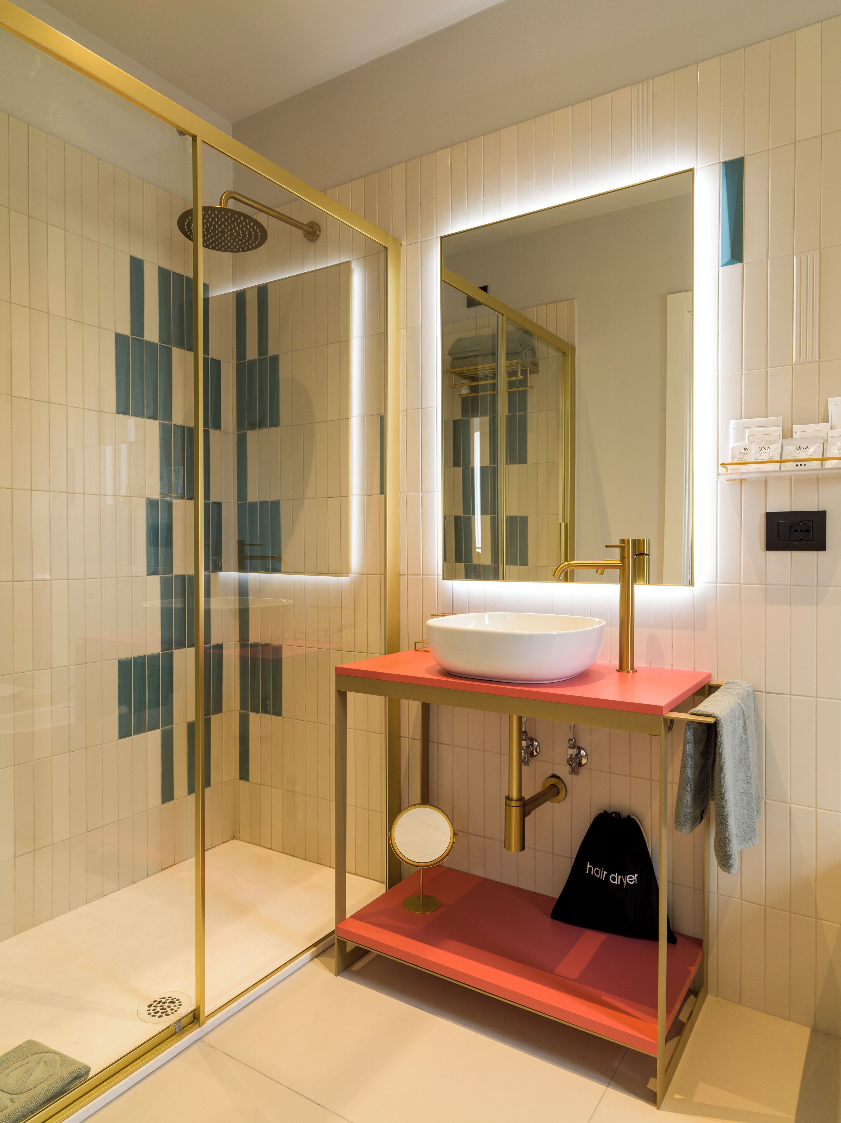 Un bagno moderno con doccia vetrata, lavandino bianco su un piano di lavoro rosso, rubinetto dorato e specchio sopra il lavandino.