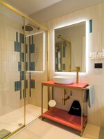 Un bagno moderno con doccia vetrata, lavandino bianco su un piano di lavoro rosso, rubinetto dorato e specchio sopra il lavandino.