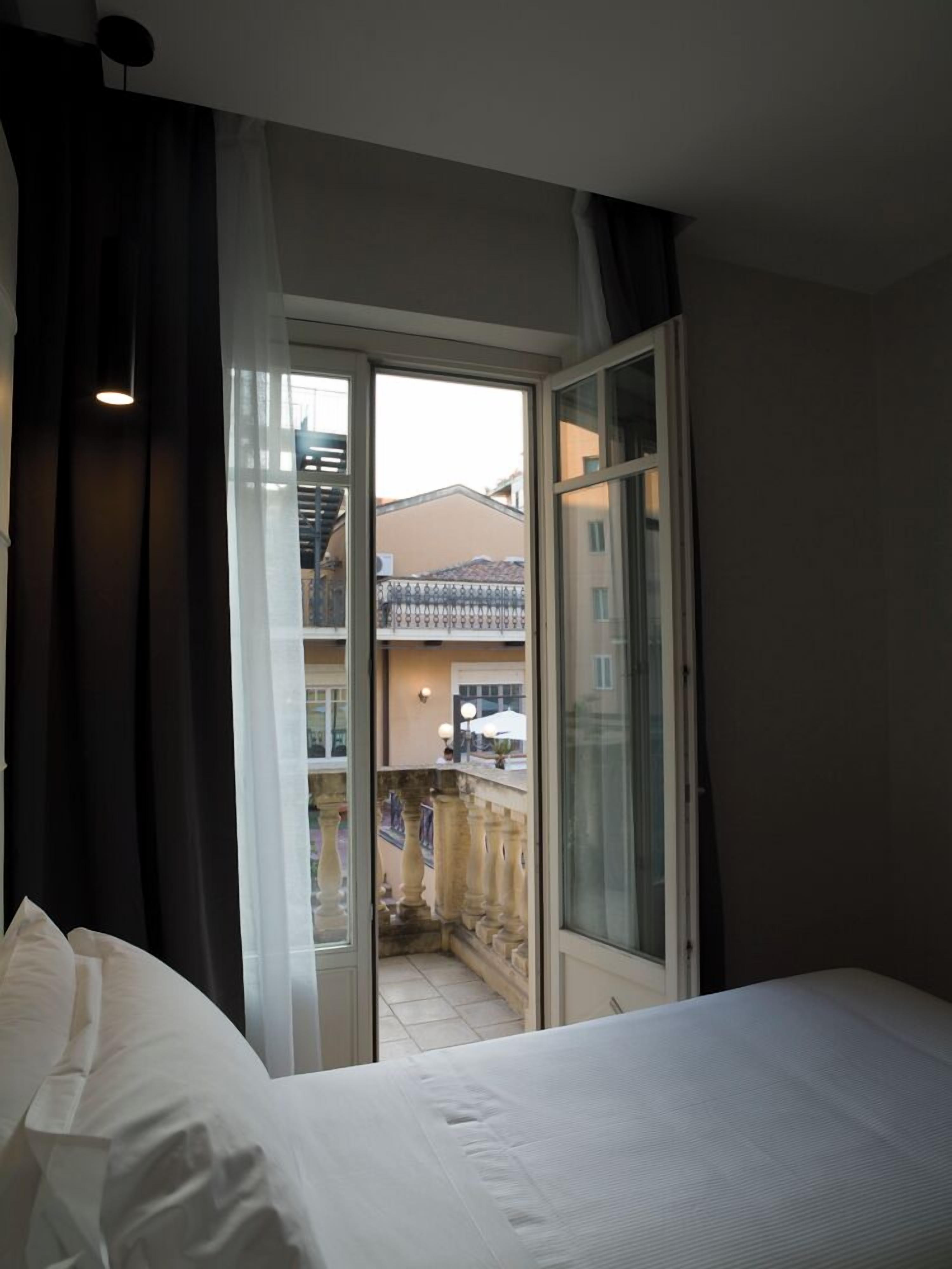 A hotel room with a bed, a balcony with a view of buildings, and a door leading to the balcony.