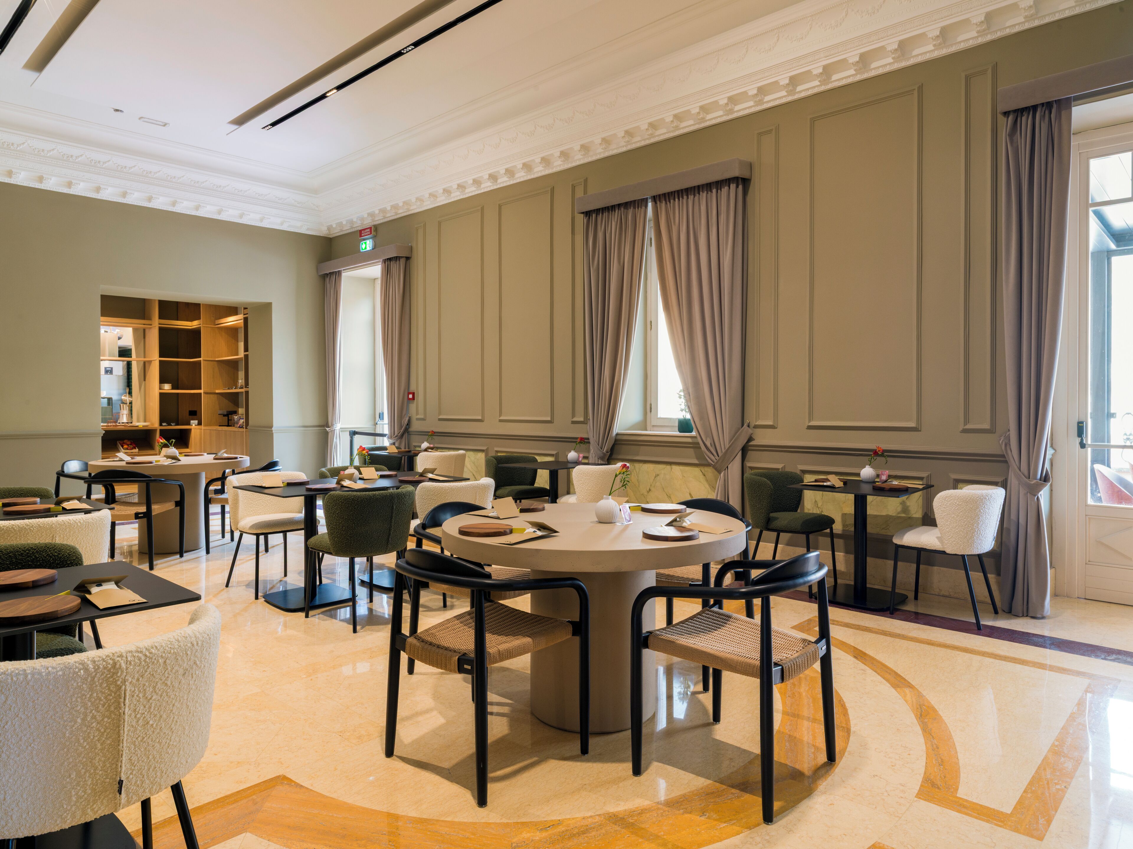 A spacious dining area with round tables and chairs, a built-in bookshelf, and large windows with curtains.