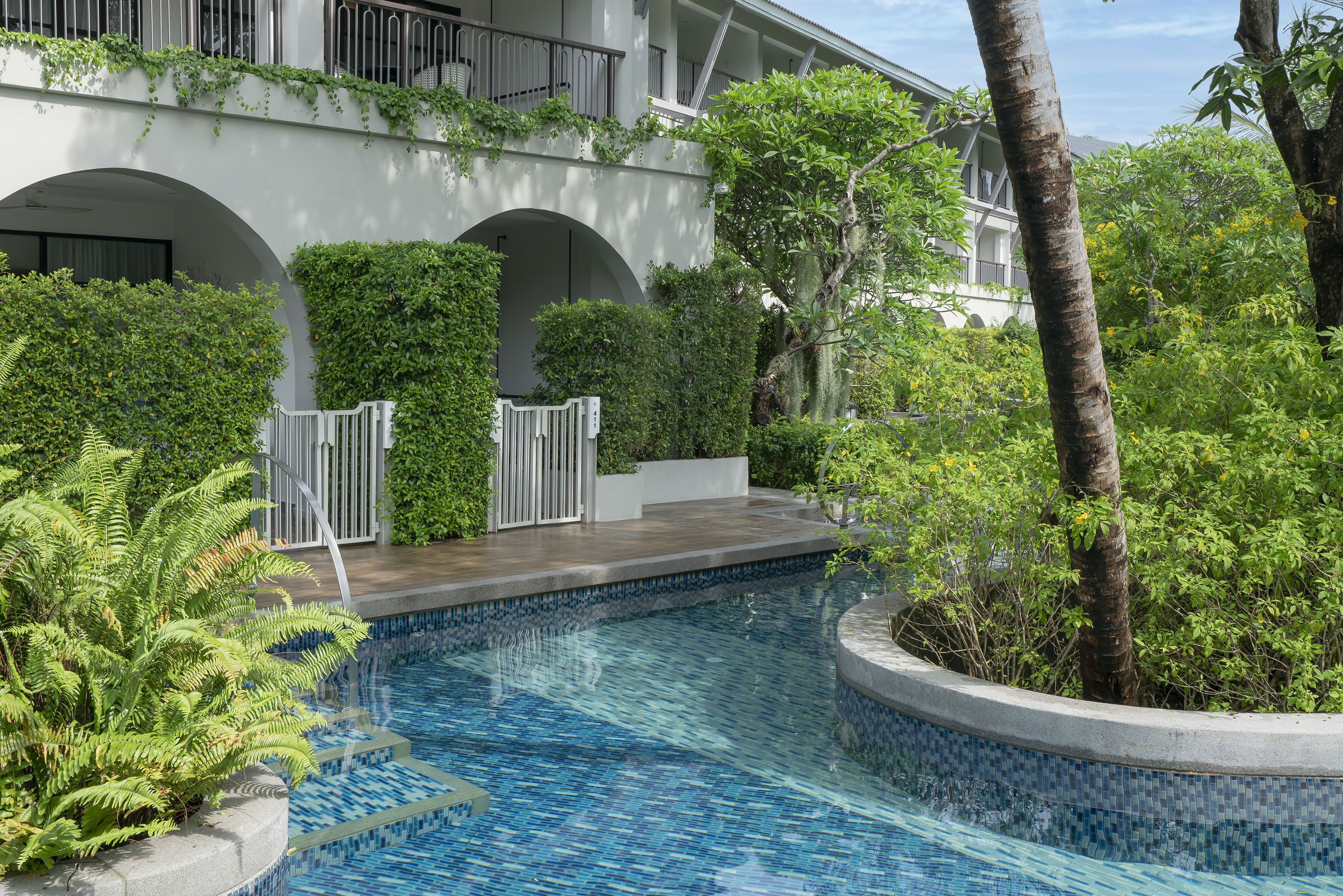 A swimming pool with a tiled edge, surrounded by lush greenery and a white building with balconies.