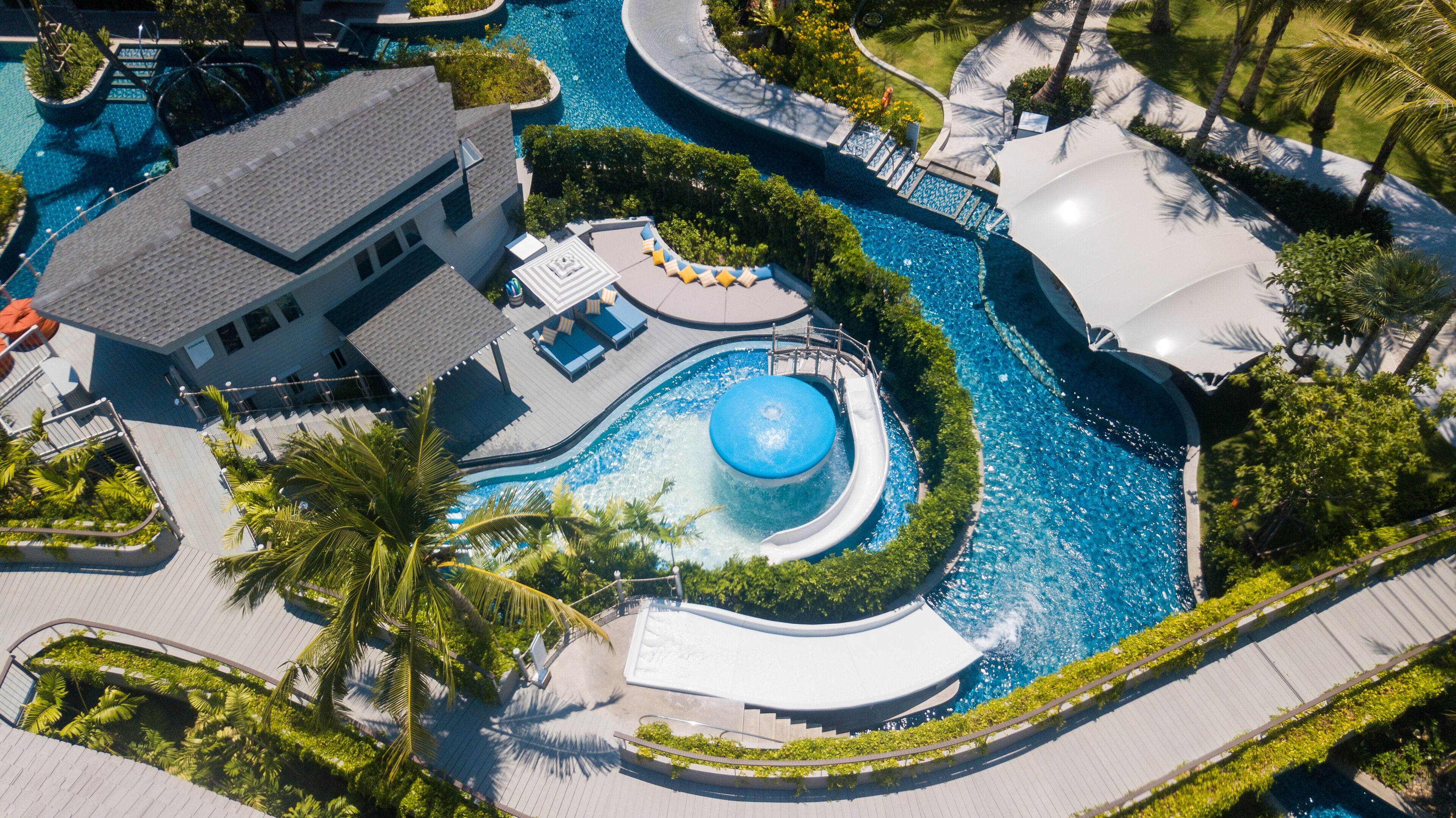 An aerial view of a resort with a large central pool, a circular water feature, and surrounding palm trees.