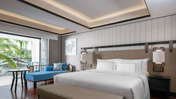 A modern hotel room with a large bed, a seating area with a blue sofa, a small table, and a balcony with a view of palm trees.