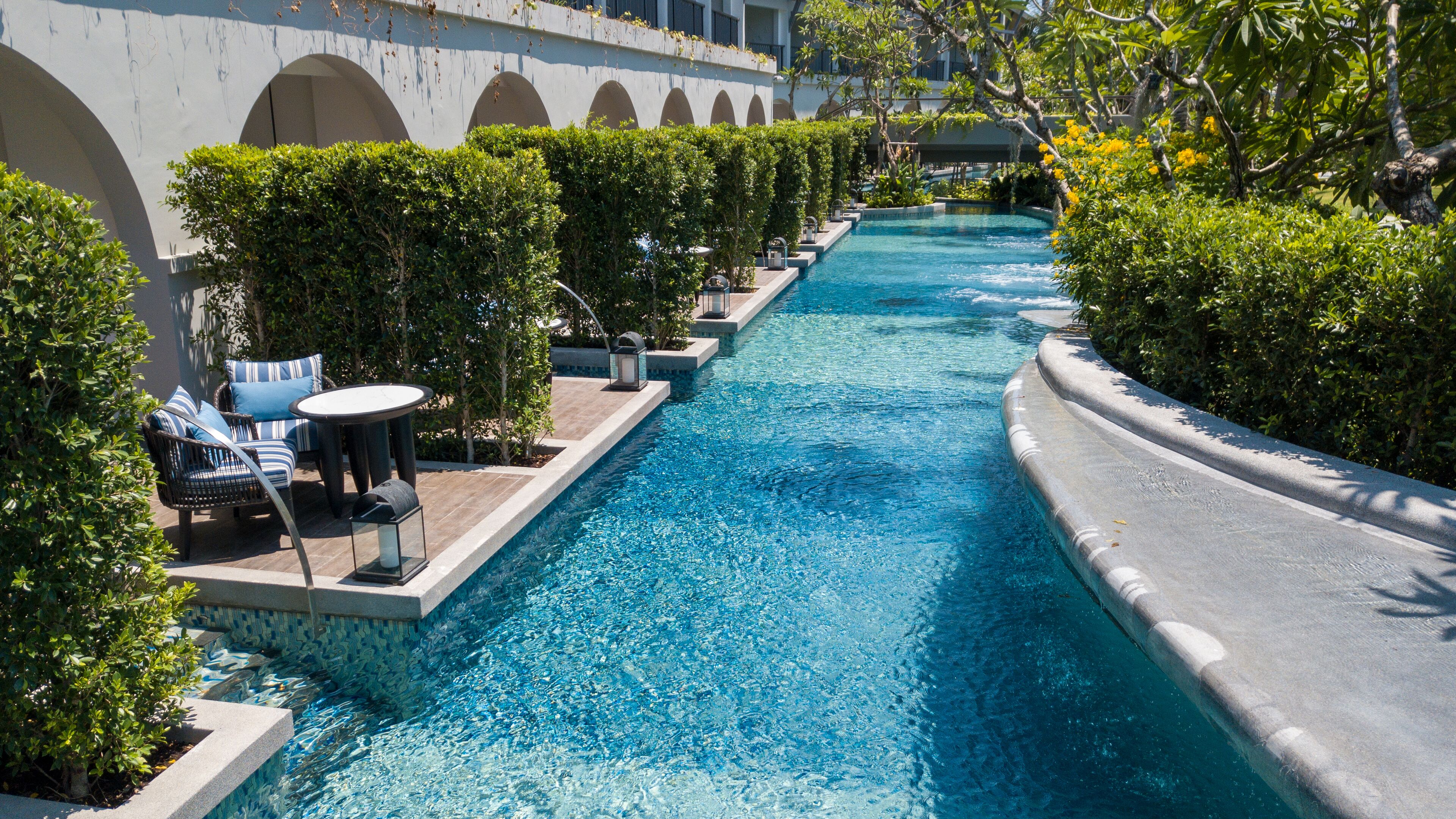 A long, narrow swimming pool with clear blue water, surrounded by neatly trimmed hedges and a row of lounge chairs and tables.