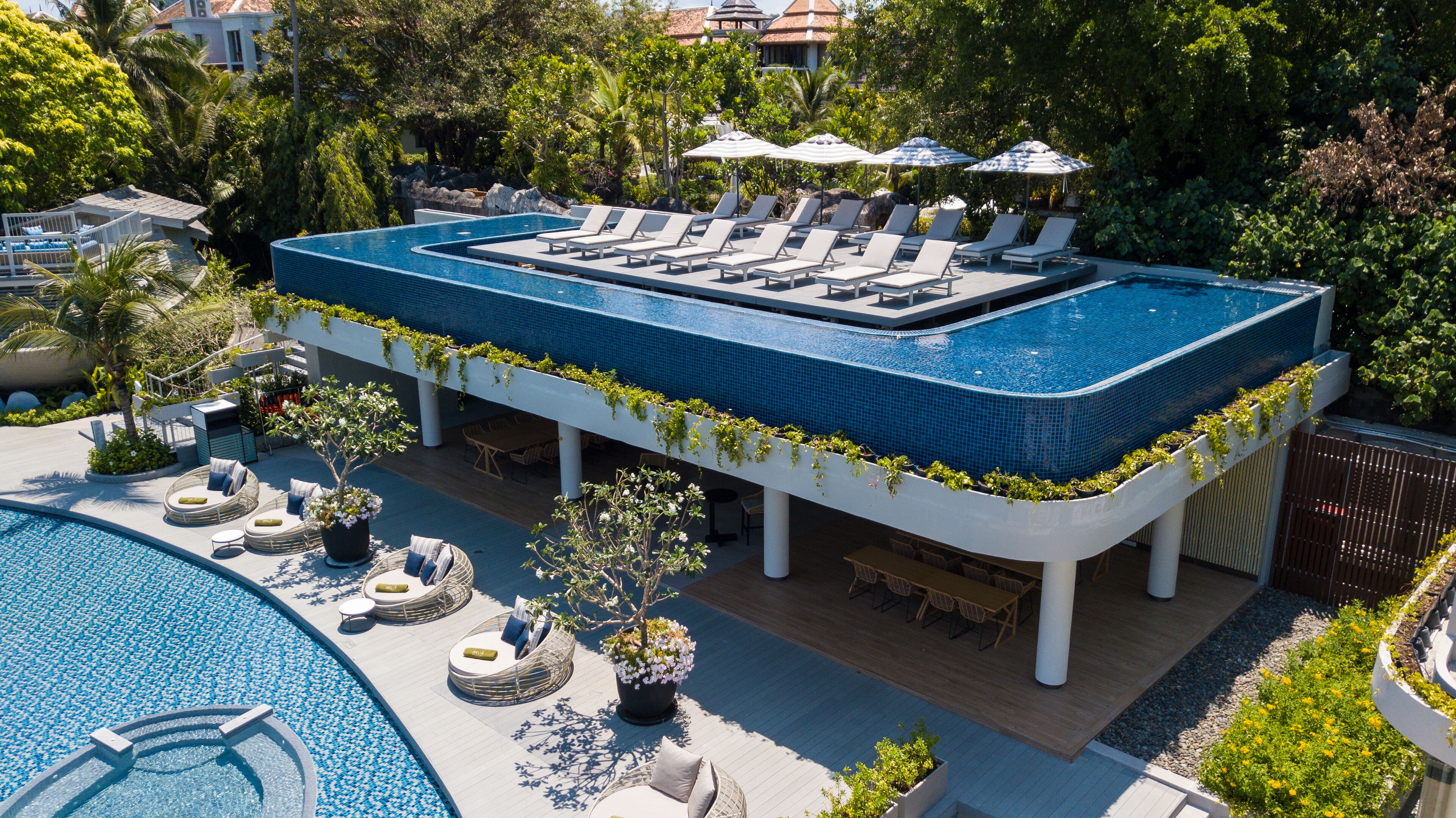 A rooftop infinity pool with lounge chairs and umbrellas, surrounded by lush greenery and a view of a residential area.