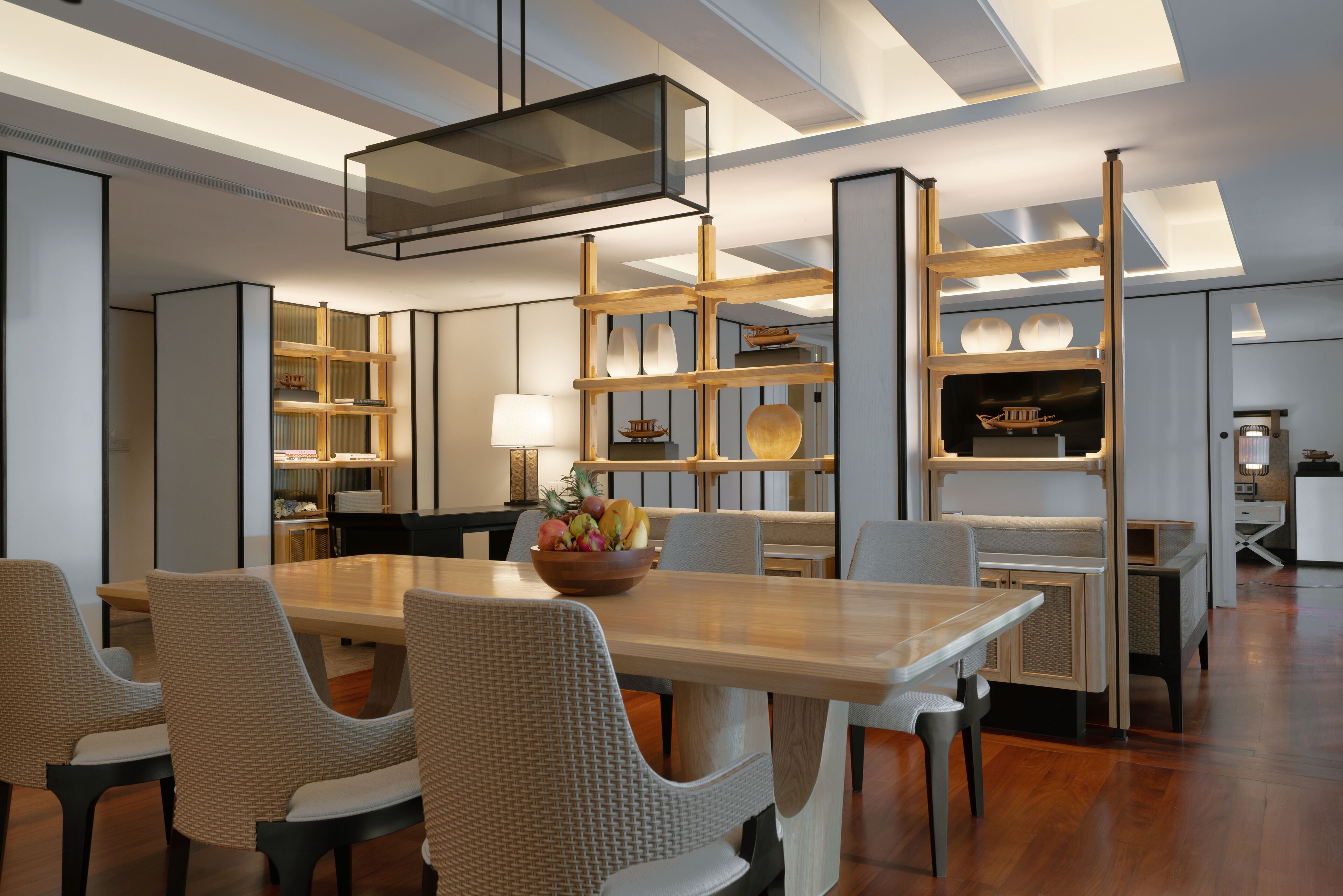 A modern dining area with a wooden table, wicker chairs, and a fruit bowl. There are built-in shelves with decorative items and a lamp.