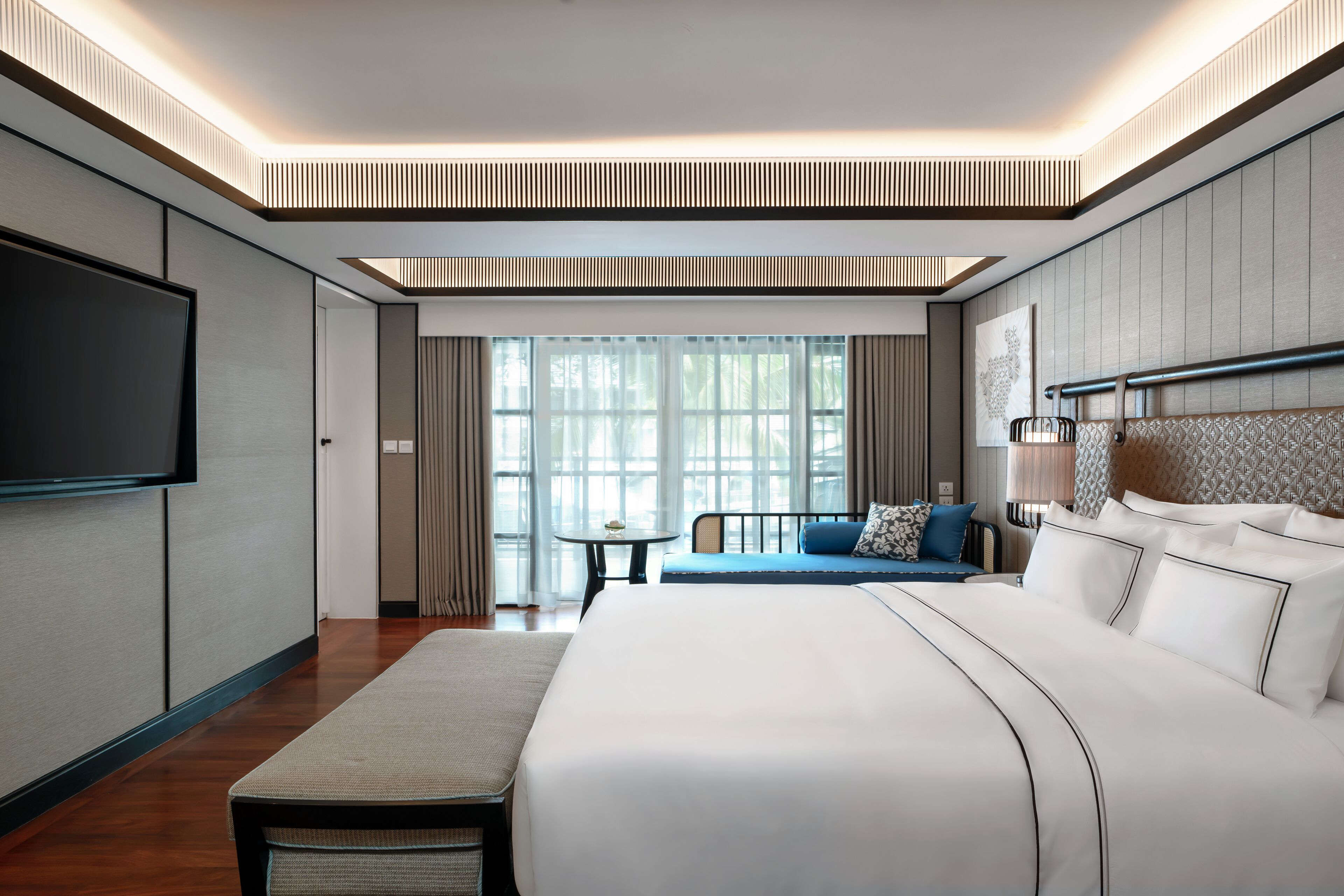 A modern hotel room with a large bed, a seating area, a flat-screen TV, and large windows.