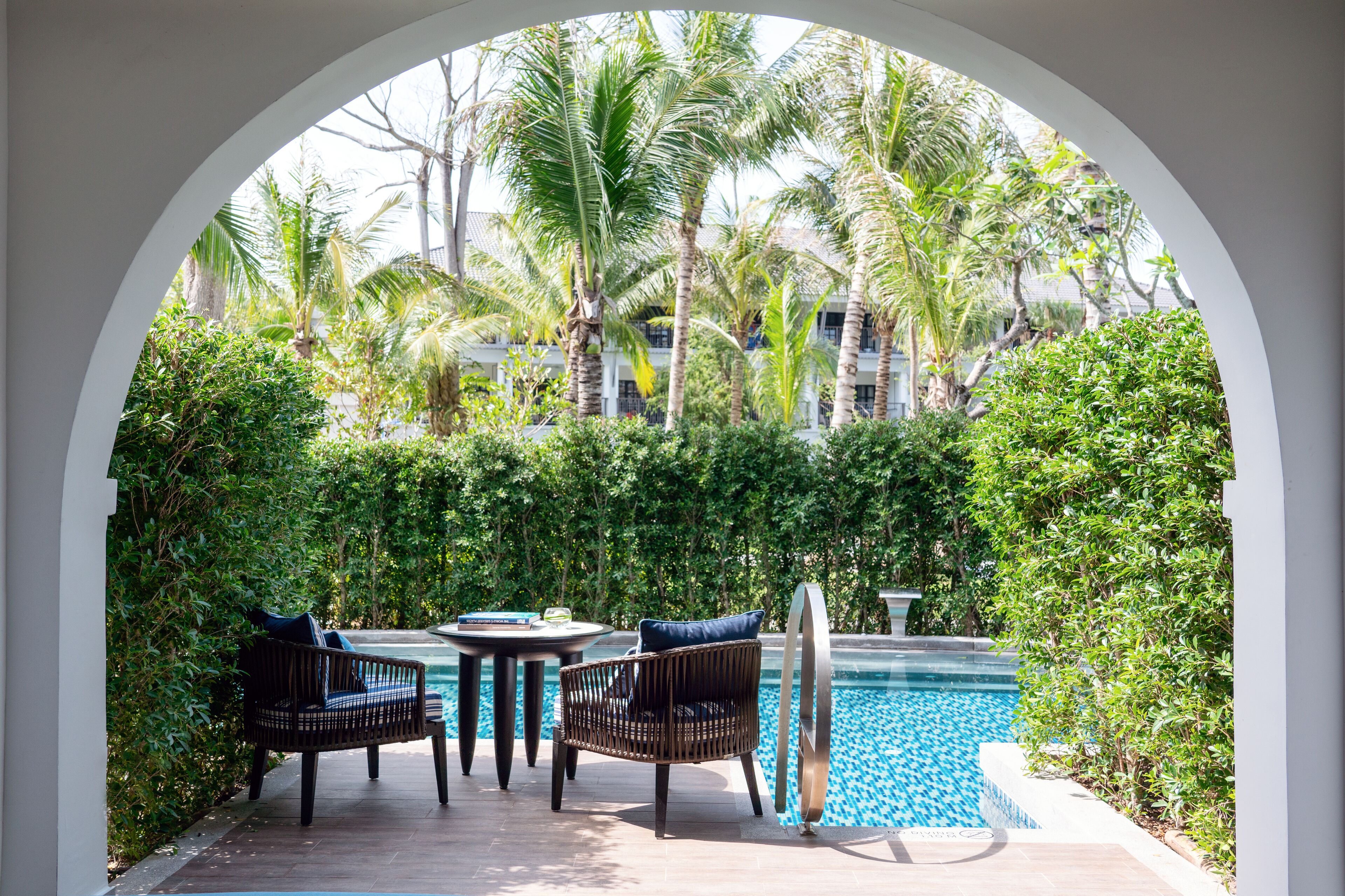 An outdoor seating area with a pool, surrounded by lush greenery and palm trees.
