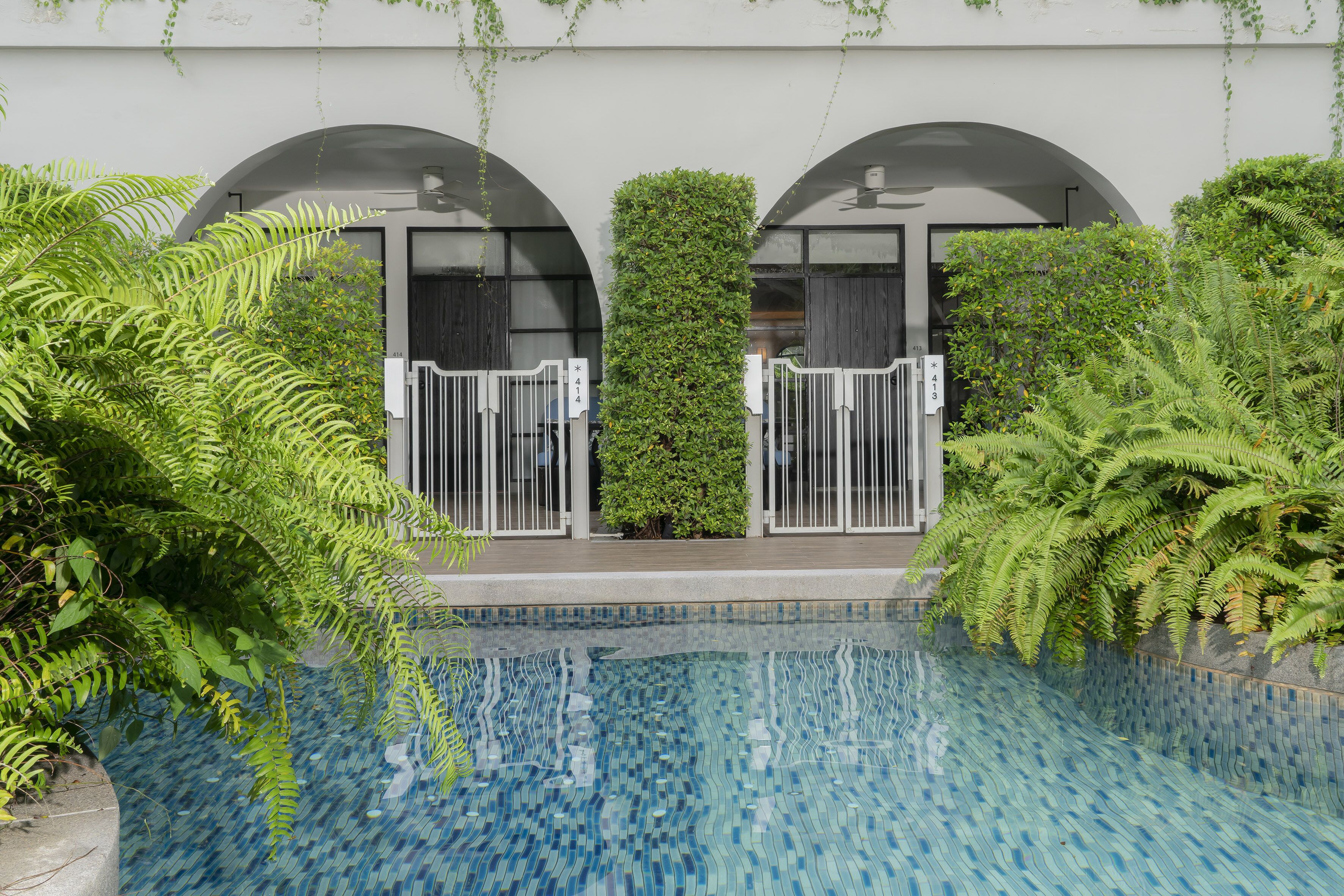 A swimming pool with a tiled edge, surrounded by lush greenery and a building with arched windows.