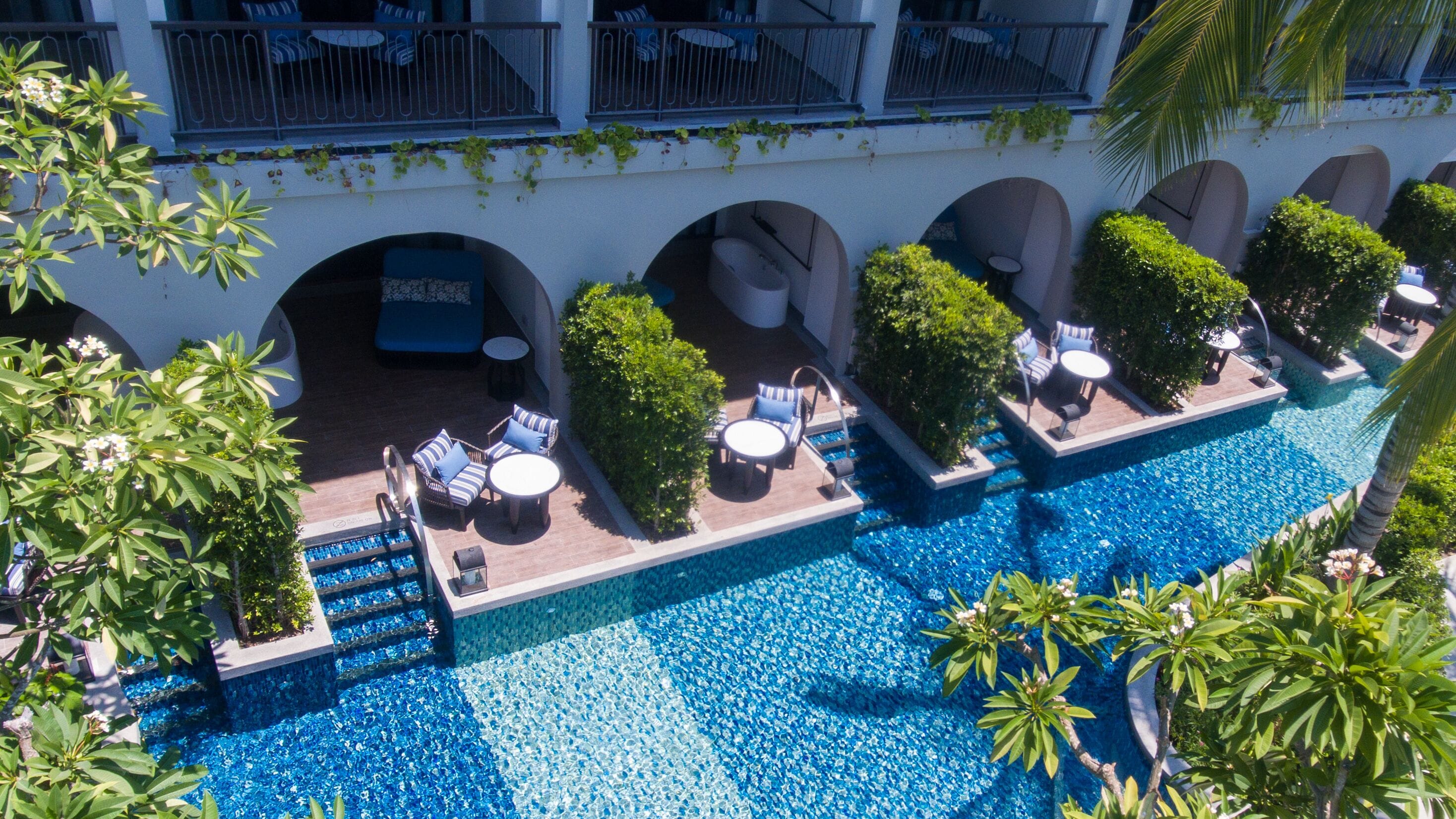 A rooftop pool area with lounge chairs and a small seating area, surrounded by greenery and a white building facade.