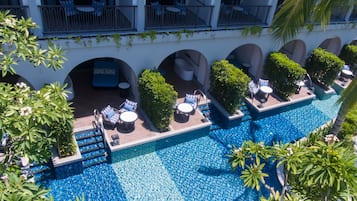 A rooftop pool area with lounge chairs and a small seating area, surrounded by greenery and a white building facade.