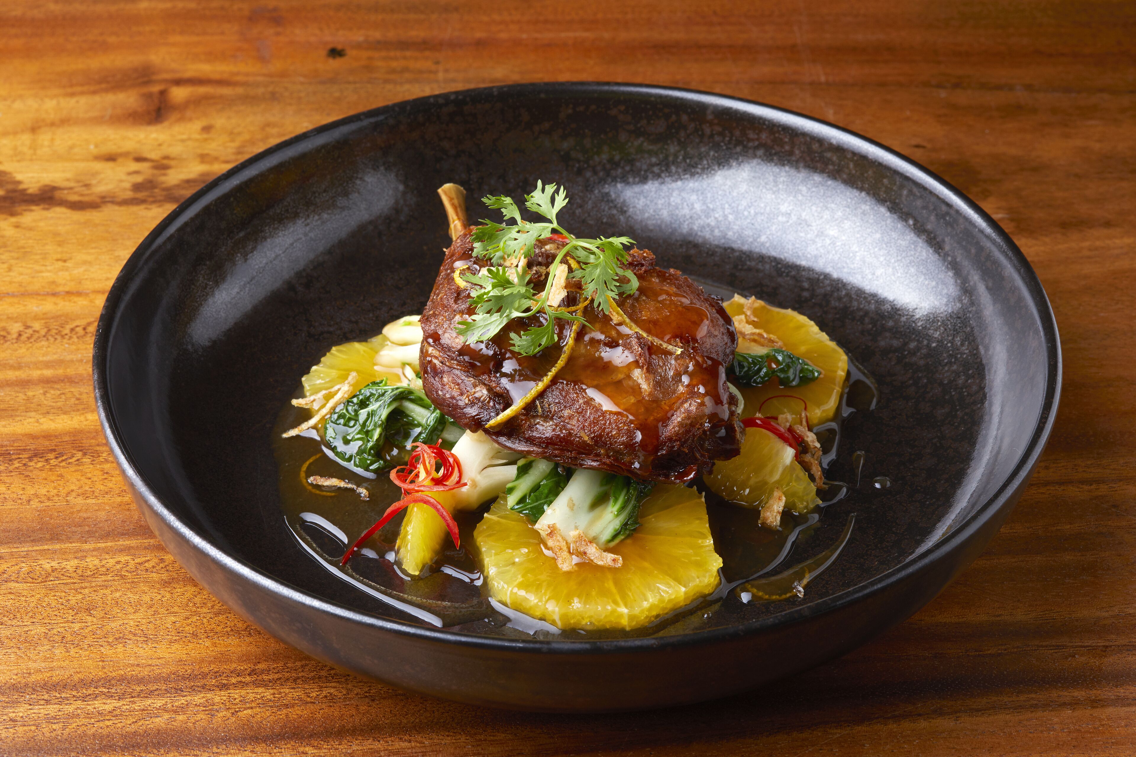 A bowl of food with a piece of meat, vegetables, and garnishes on a wooden table.