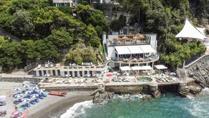 An aerial view of a coastal hotel with a pool, beach umbrellas, and a rocky shoreline.