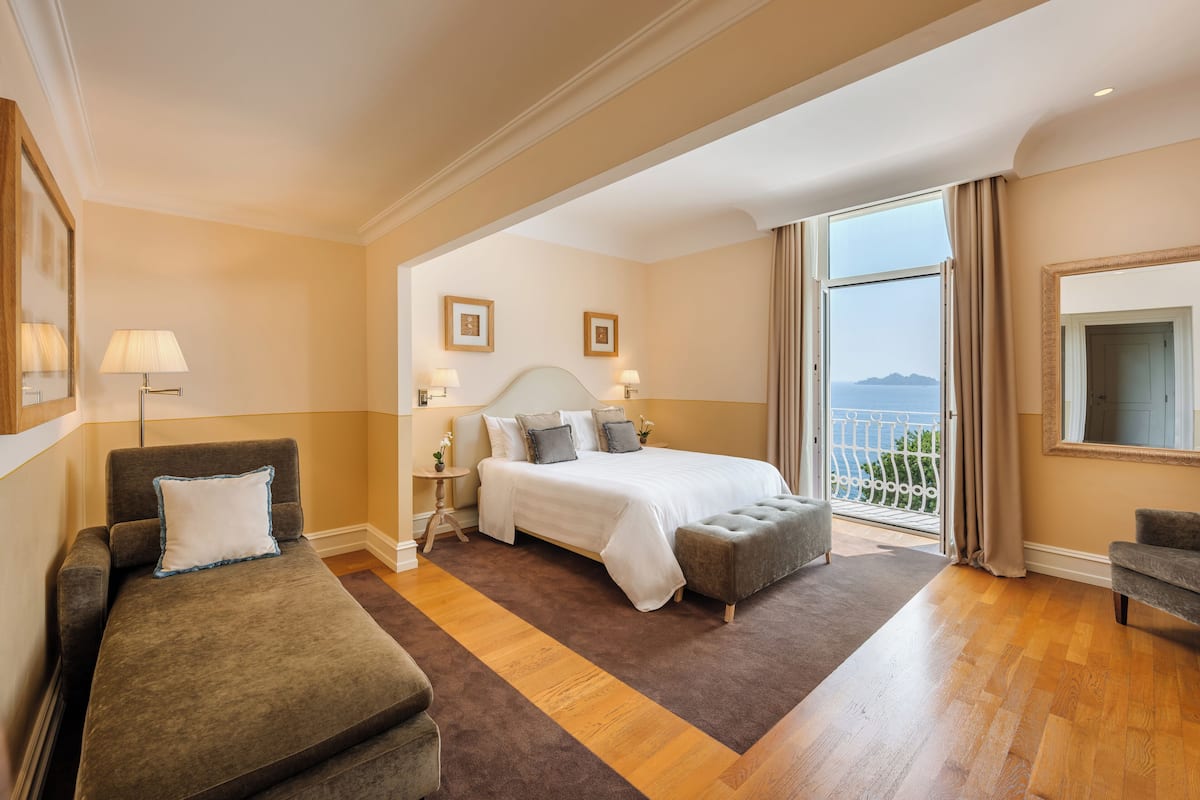 A hotel room with a bed, a sofa, an ottoman, a nightstand, and a view of the sea.