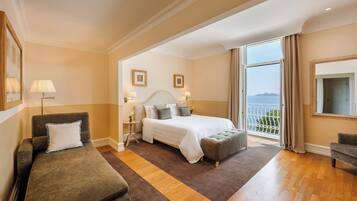 A hotel room with a bed, a sofa, an ottoman, a nightstand, and a view of the sea.