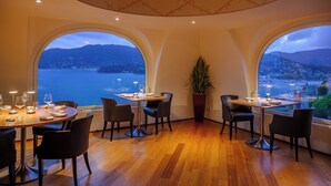 Un comedor con una mesa dispuesta para dos, ventanas amplias con vista al mar y colinas, y una planta en maceta en el centro.