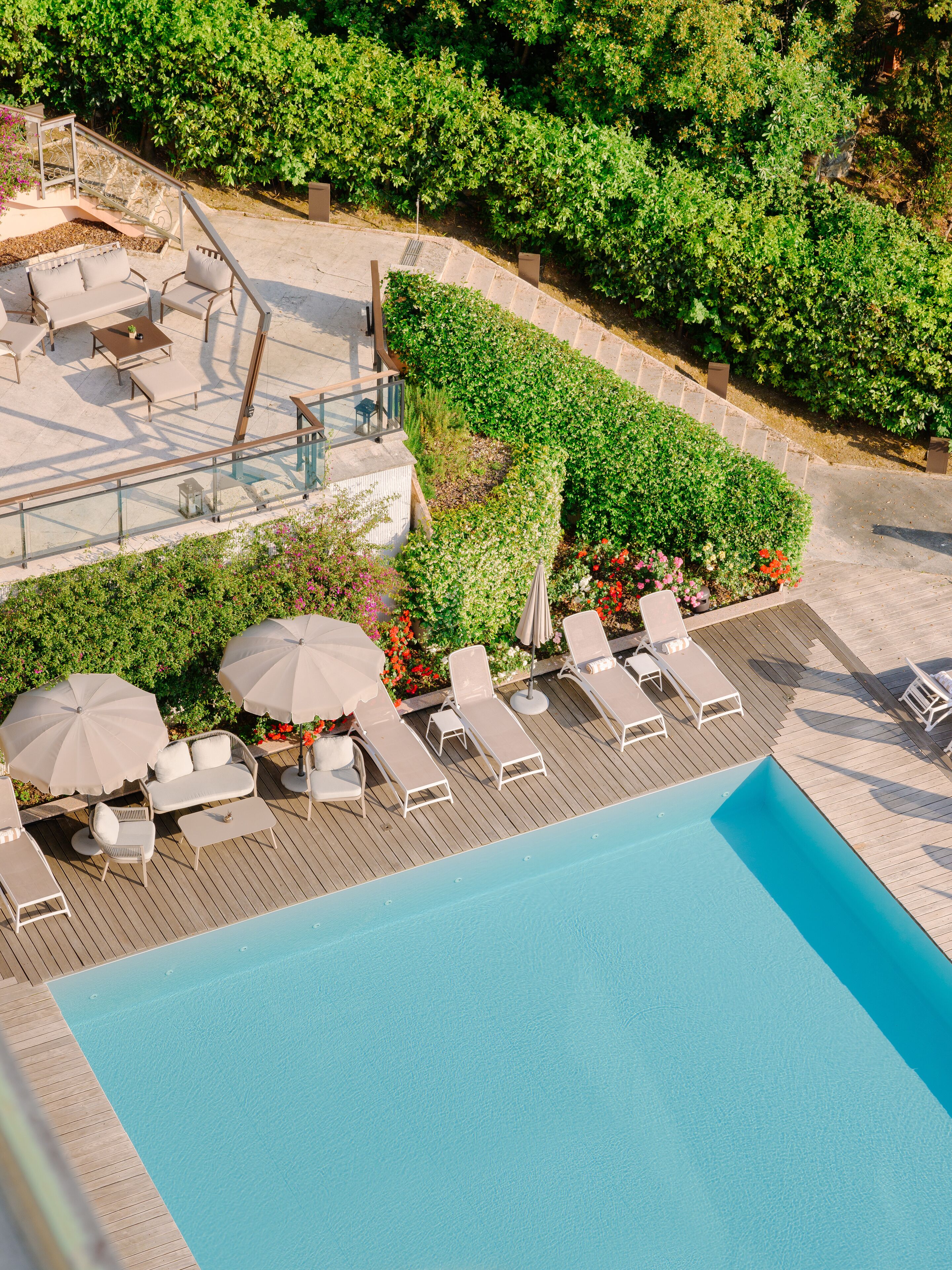Zona de piscina al aire libre con sillones de descanso y sombrillas, rodeada de follaje y una terraza de madera.