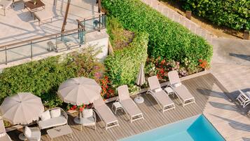 Zona de piscina al aire libre con sillones de descanso y sombrillas, rodeada de follaje y una terraza de madera.