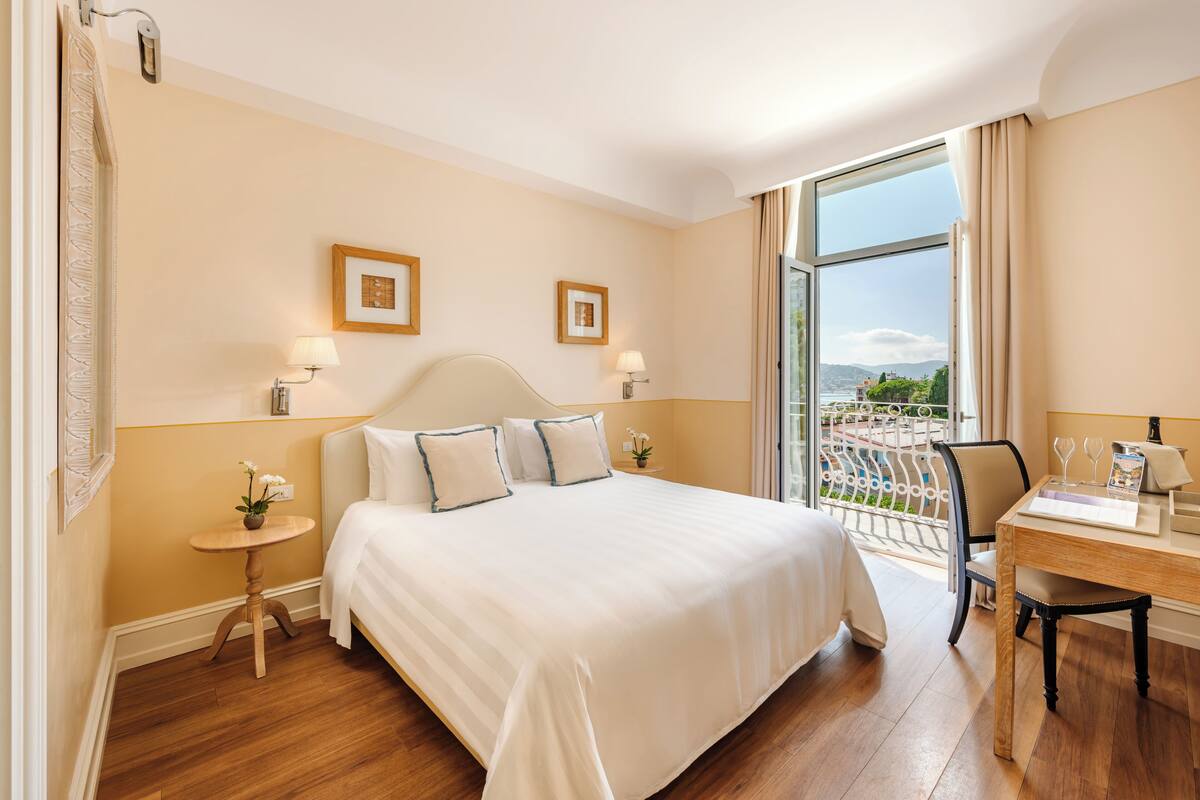 A hotel room with a large bed, a desk with a chair, a small table with a lamp, and a view of the outdoors through a large window.