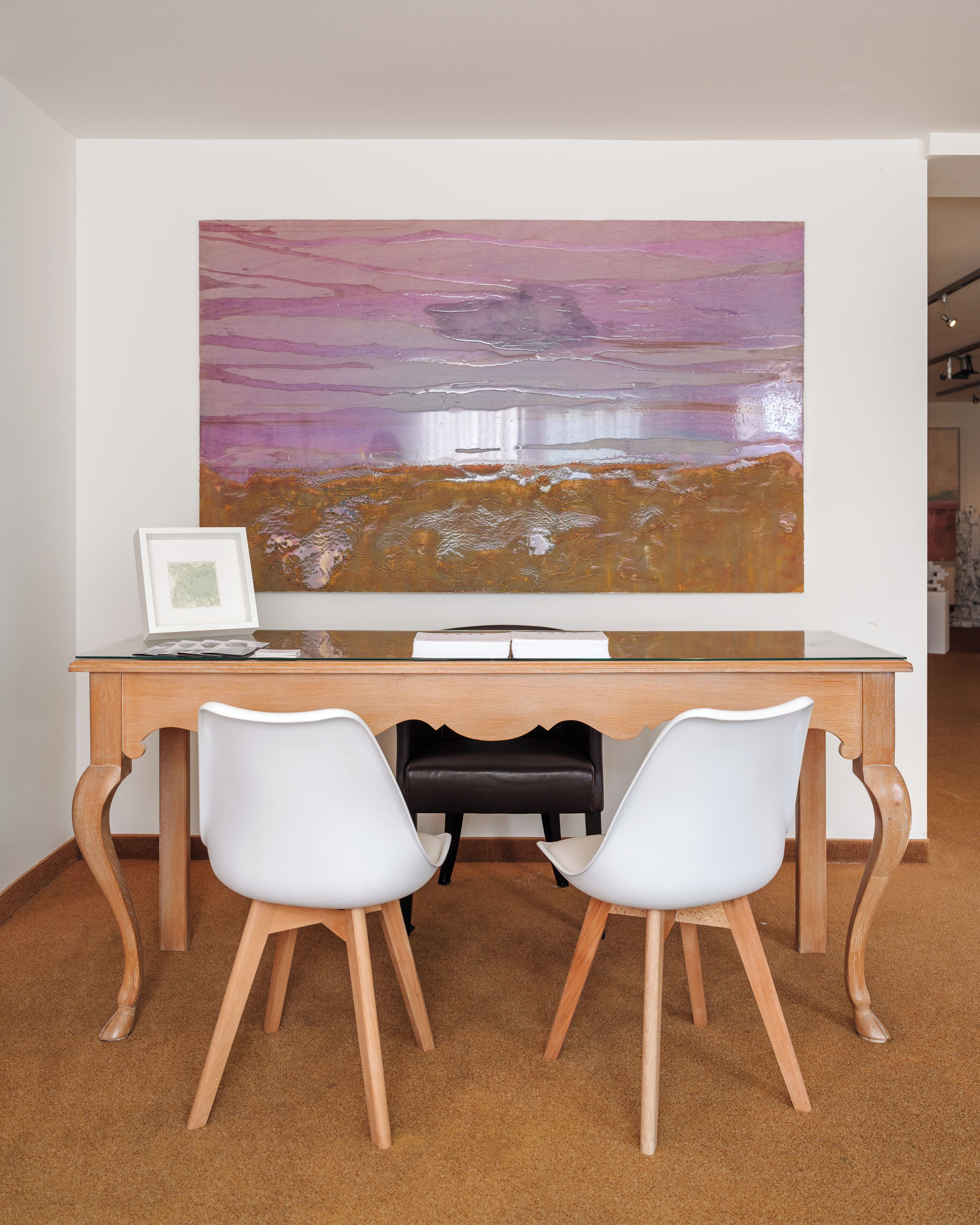 A desk with two white chairs, a large abstract painting, and a framed picture on the wall.