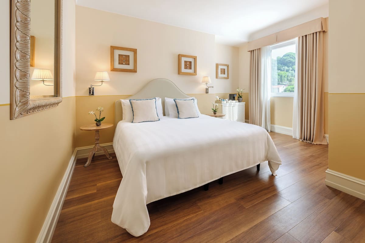 A neatly arranged bedroom with a large bed, bedside lamps, framed pictures on the wall, and a view of greenery outside.