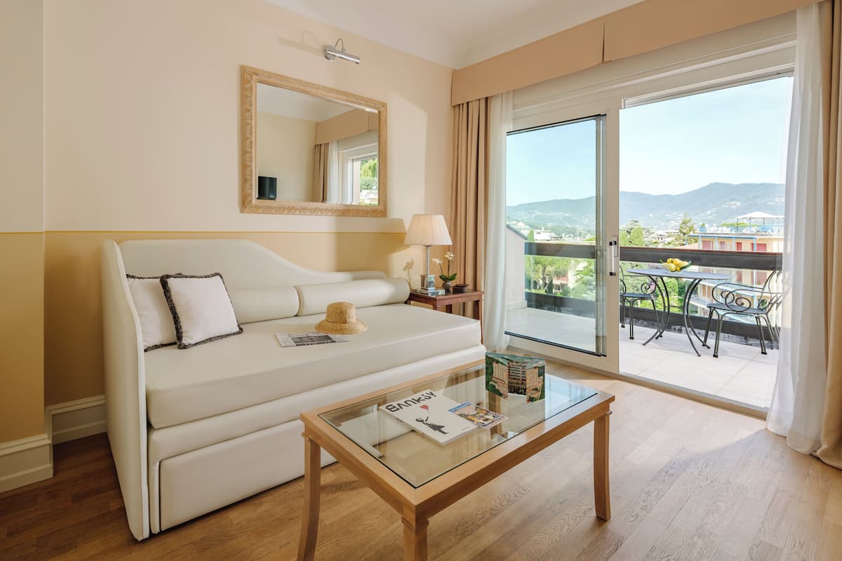 A hotel room with a sofa, a glass coffee table, a lamp, a mirror, and a balcony with outdoor furniture.