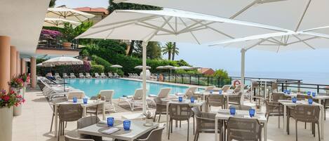 A poolside area with multiple tables and chairs, umbrellas, and a view of the sea.
