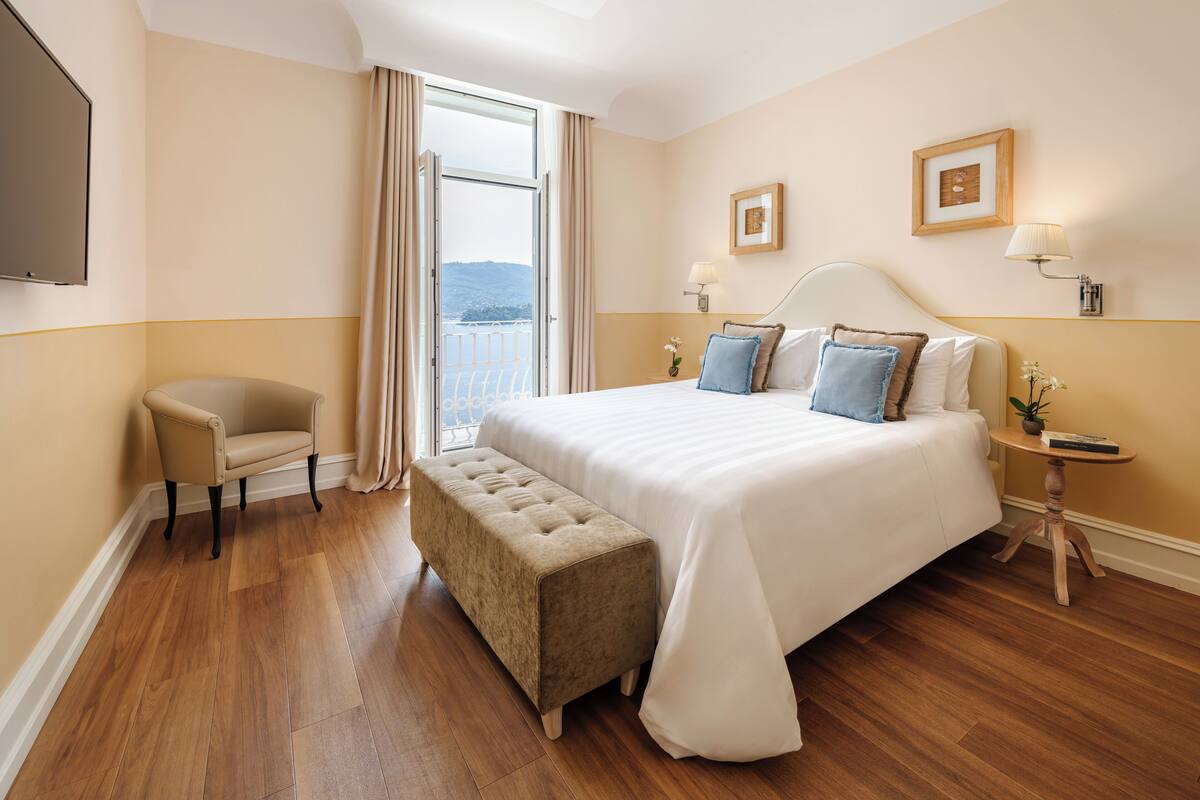 A hotel room with a large bed, a chair, a bench, a nightstand with a lamp, and a view of the outdoors.
