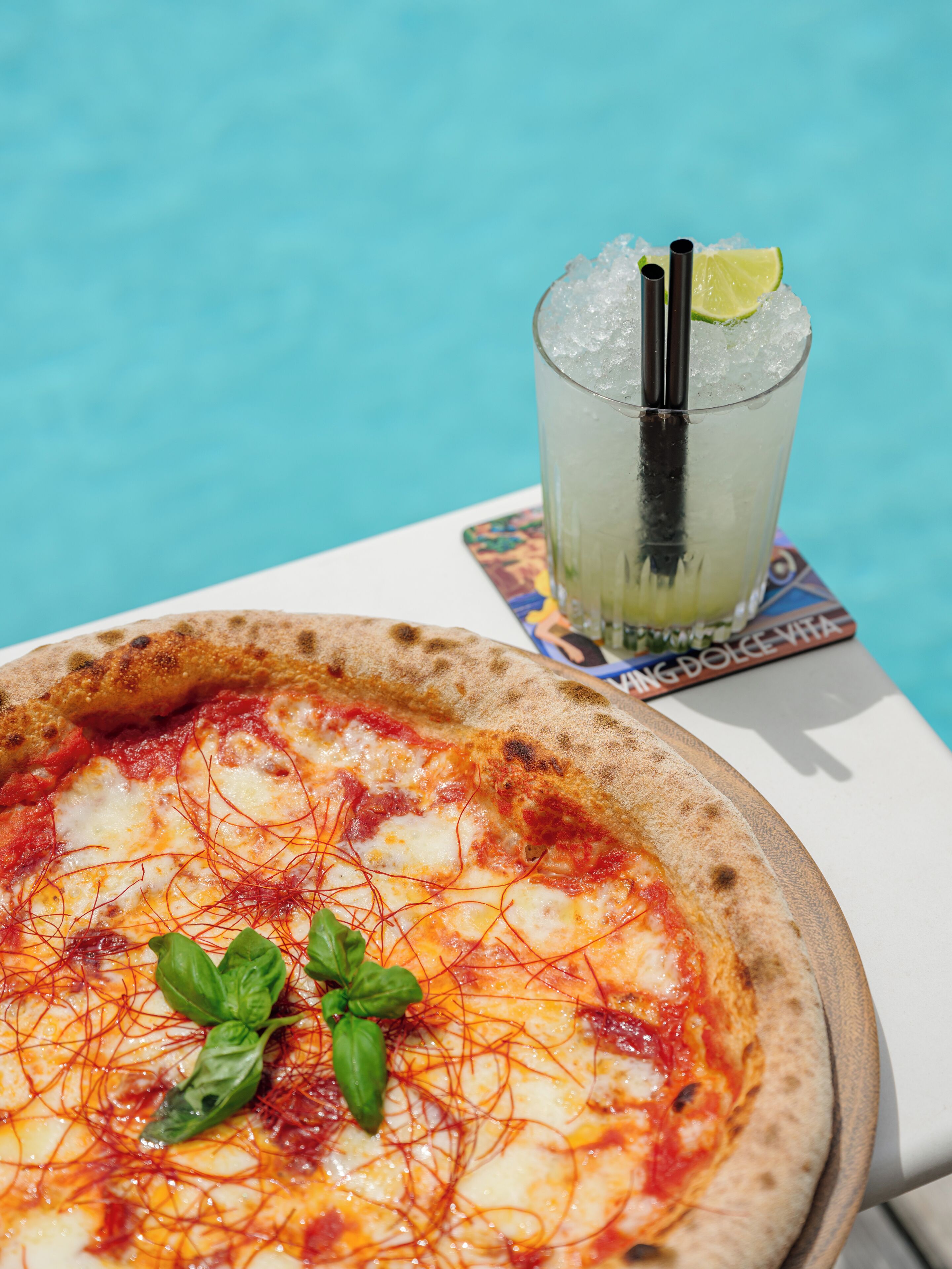 Pizza mit Basilikumblättern und ein Glas Eisgetränk mit Strohhalm und Limettenscheibe.