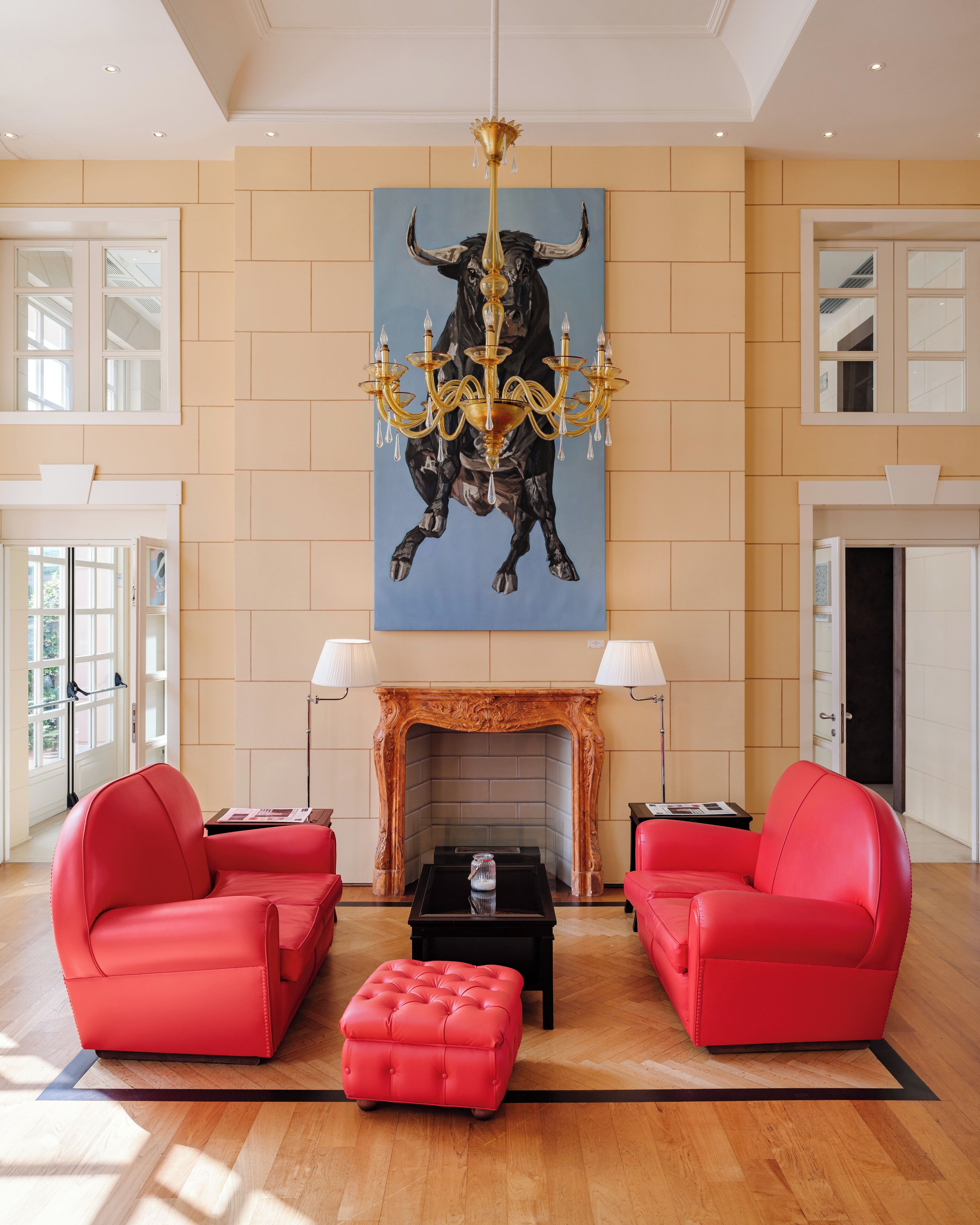Un vaste salon agrémenté d’un grand tableau représentant un taureau, d’un lustre, d’une cheminée et de canapés en cuir rouge.