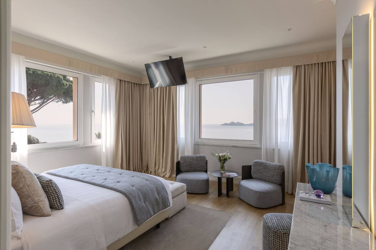 A hotel room with a bed, two armchairs, a small table with a vase, and a large window with curtains.