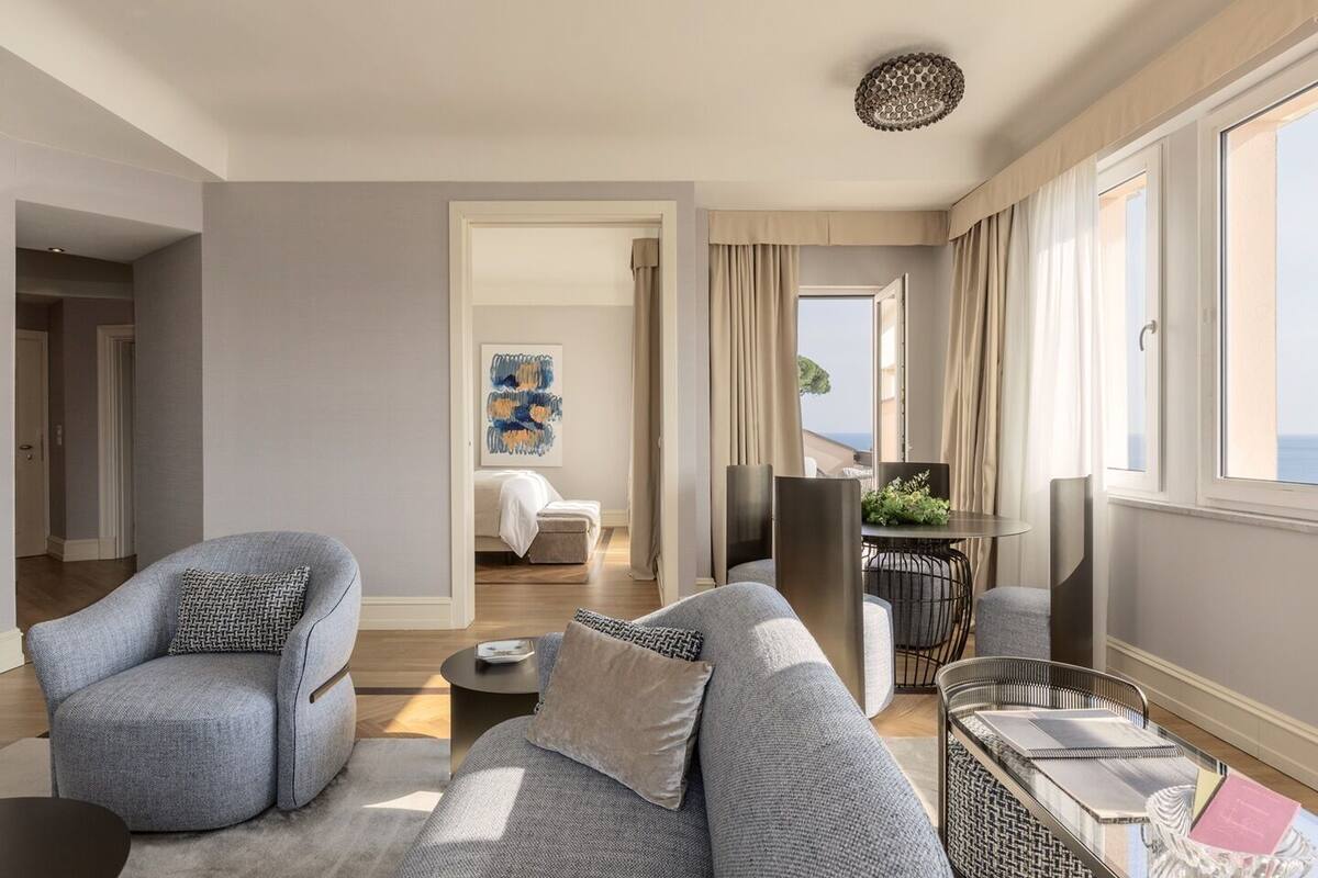 A modern living room with a sofa, armchairs, a coffee table, and a dining area with a table and chairs. There is a painting on the wall and a view of the sea through the windows.