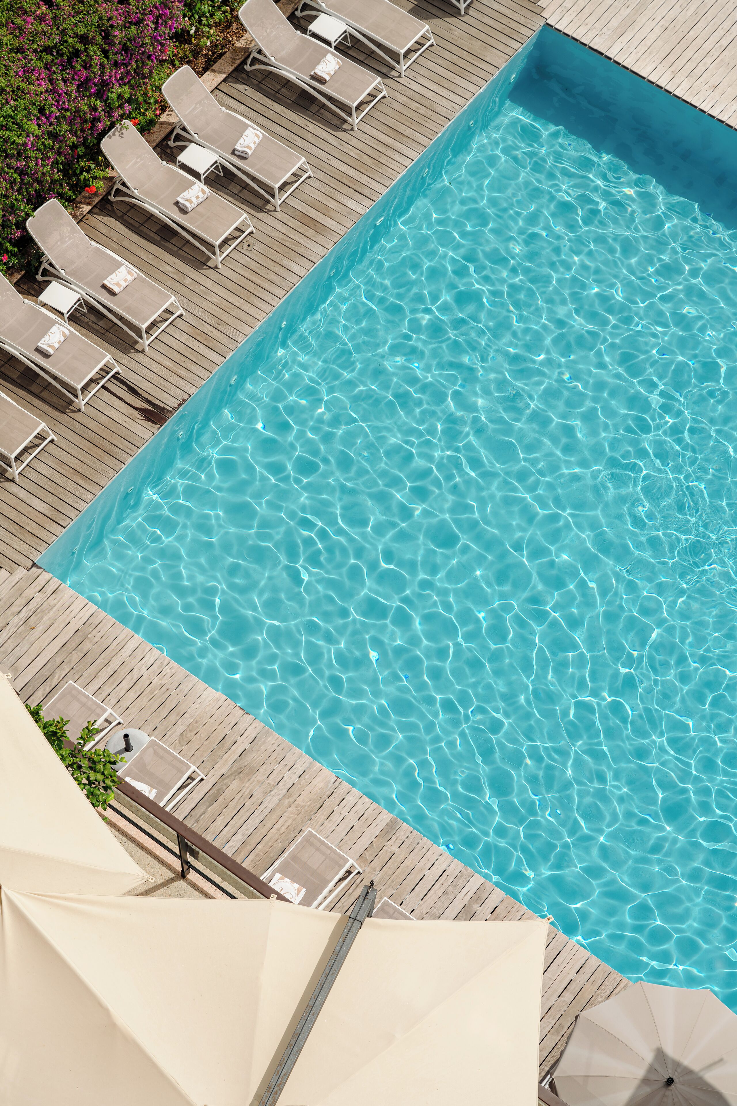 Uma piscina retangular com água azul cristalina, rodeada por um deck de madeira e espreguiçadeiras brancas.