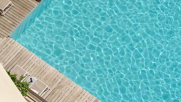 Uma piscina retangular com água azul cristalina, rodeada por um deck de madeira e espreguiçadeiras brancas.