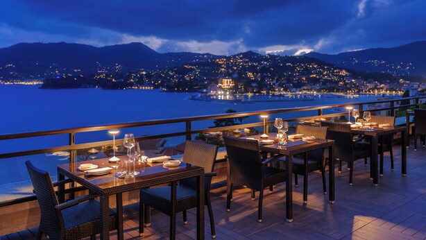 Un área de comedor al aire libre con mesas y sillas, con vistas a un cuerpo de agua y a un paisaje urbano al anochecer.