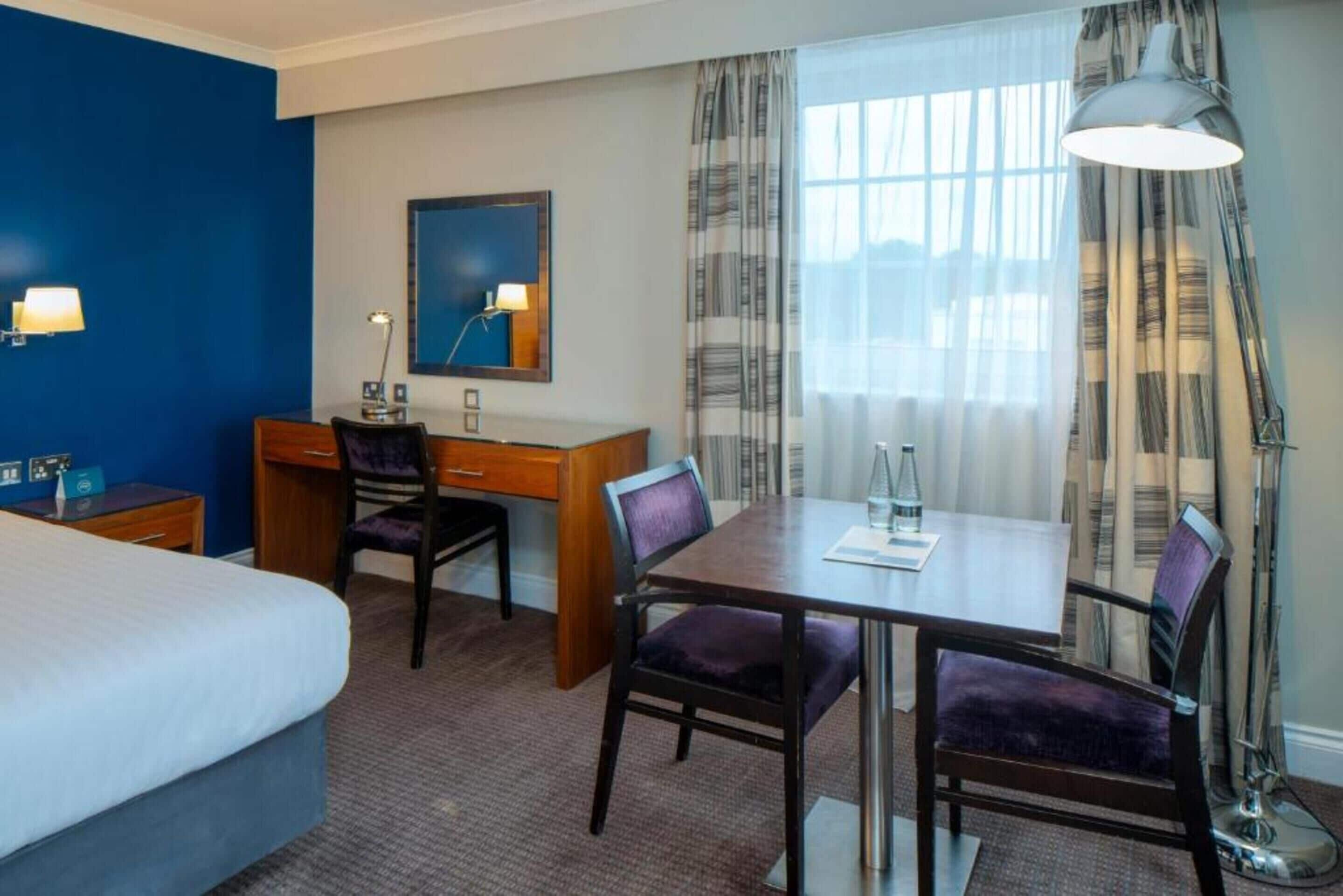 Une chambre d’hôtel avec un lit, un bureau, deux chaises, une table, une lampe, une fenêtre avec des rideaux et un tableau au mur.
